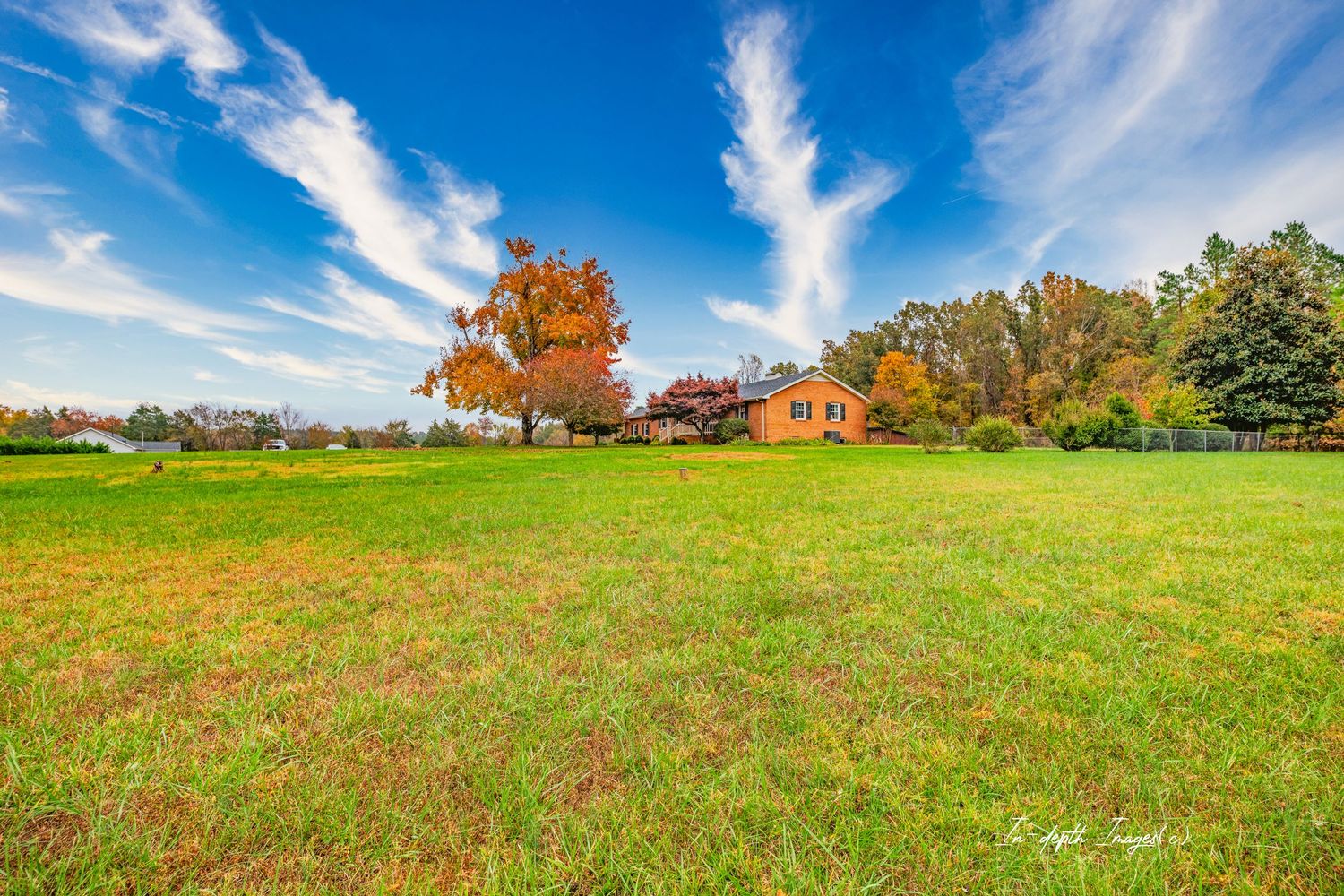 Expansive green meadow with colorful autumn trees and a brick farmhouse under a dramatic blue sky with wispy clouds.