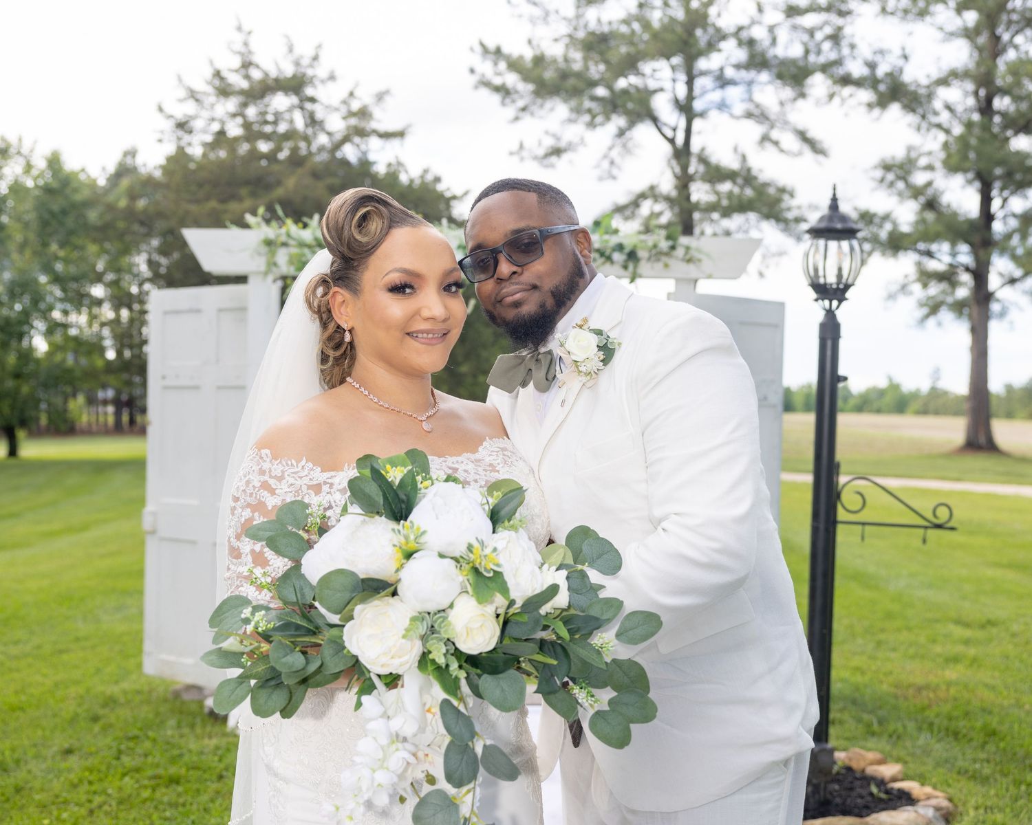 Bride and groom in white wedding attire pose together outdoors holding a white floral bouquet.