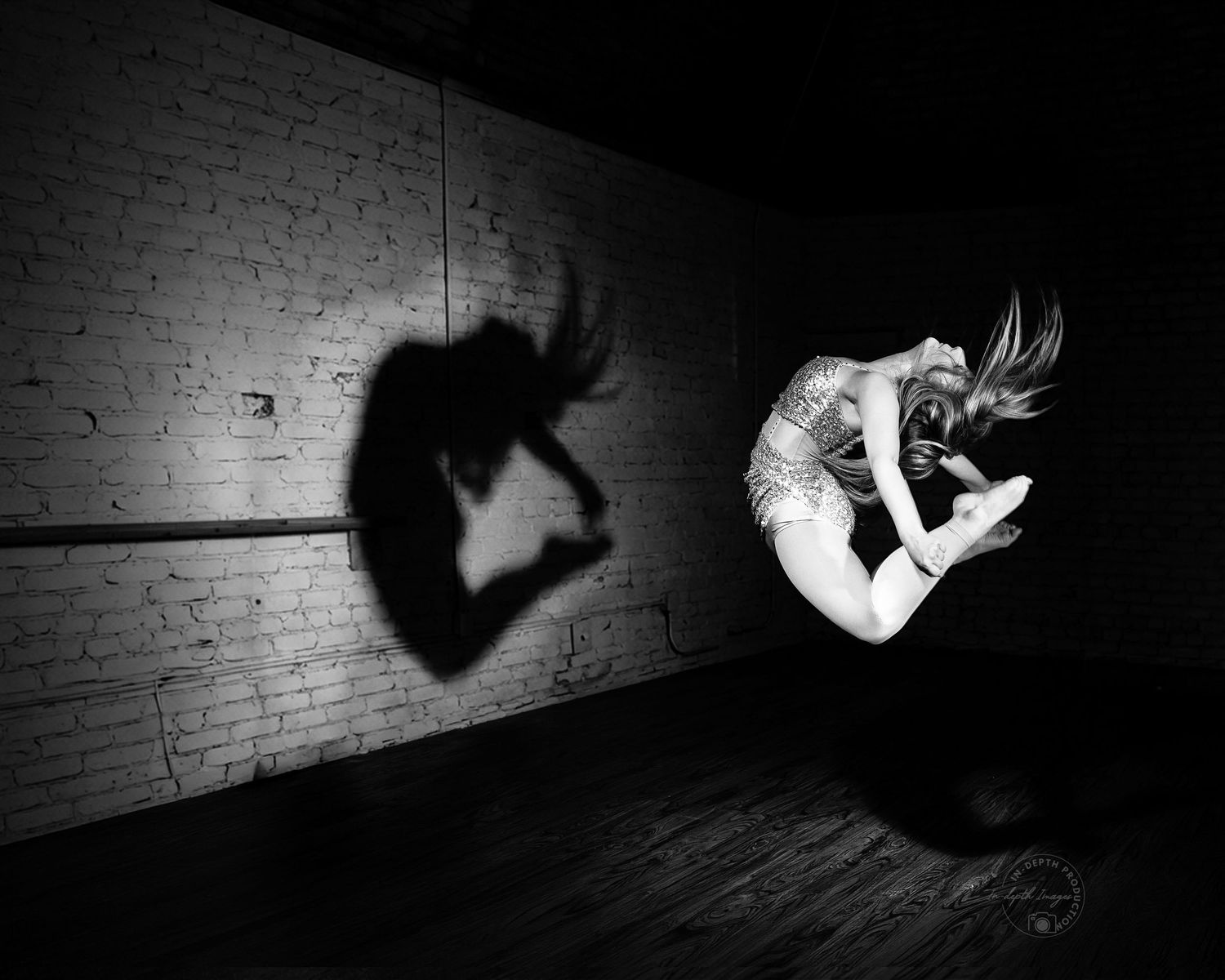 Black and white photo of a dancer leaping mid-air casting a dramatic shadow on a brick wall.