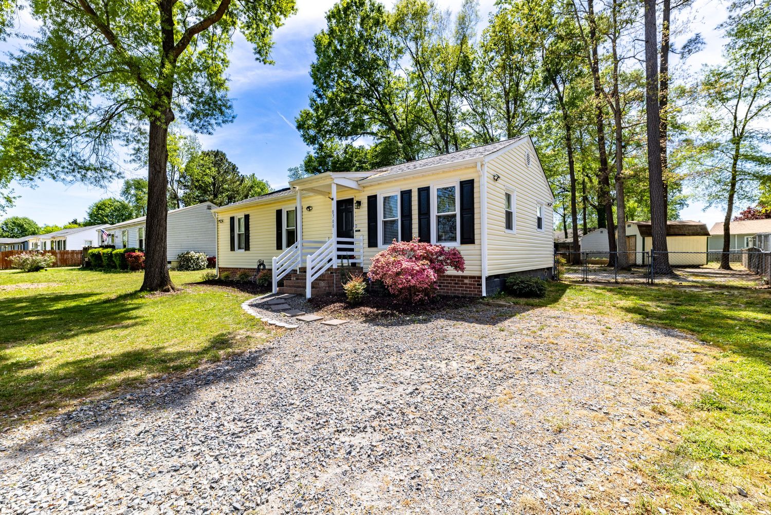 White cottage-style home with gravel driveway, blooming red bushes, and tall trees on a sunny day.