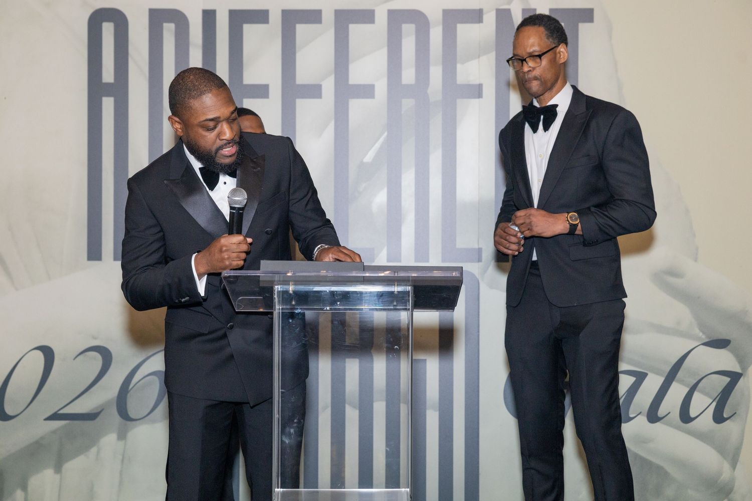Two men in tuxedos speak at a podium during a formal gala event with elegant decor in the background.