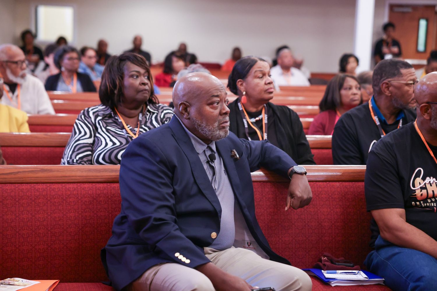 Congregation members sit in red pews during a church service, with a man in a navy blazer in the foreground and attendees behind him.