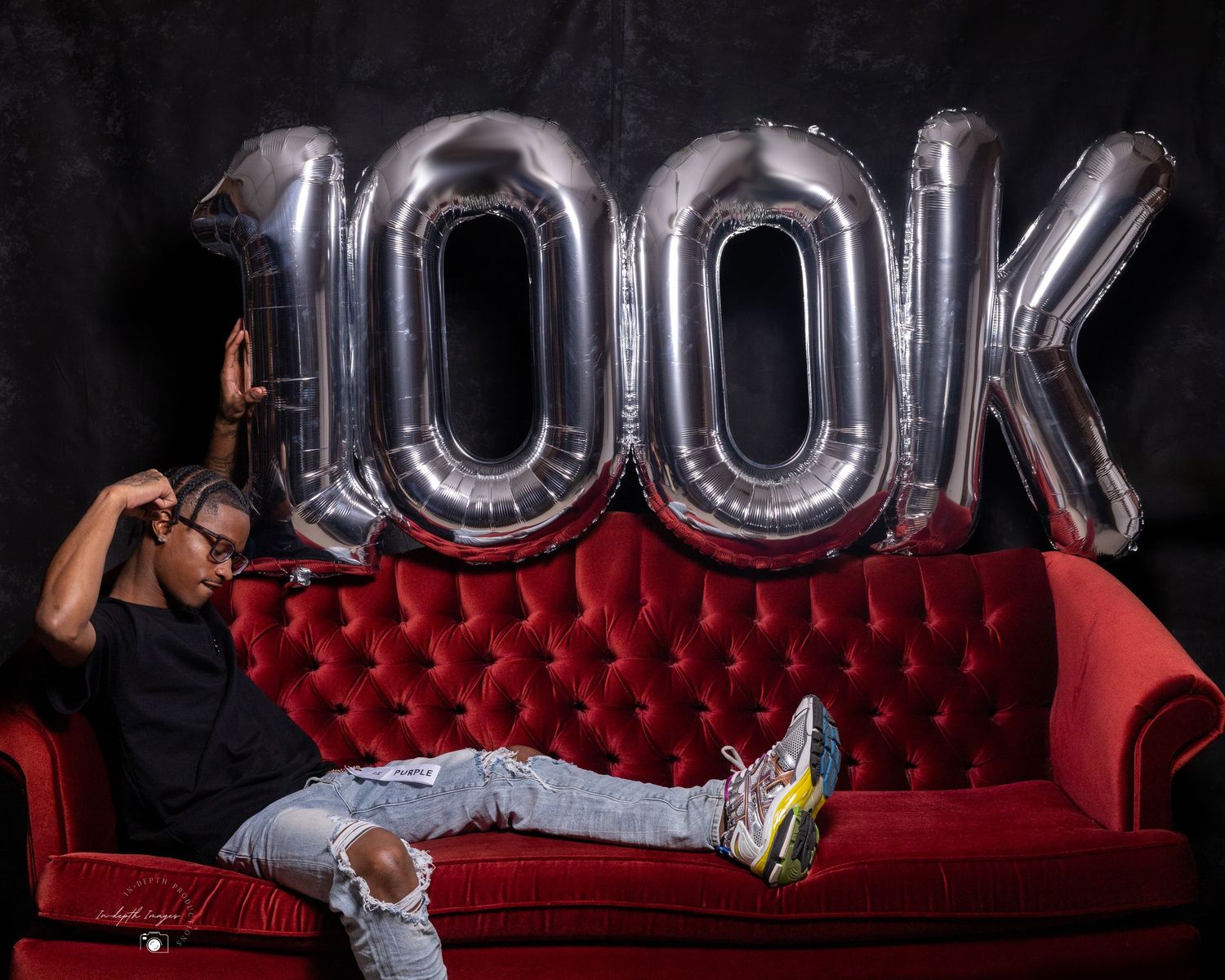 Person relaxing on red velvet sofa in front of large silver 100K balloon milestone celebration backdrop.