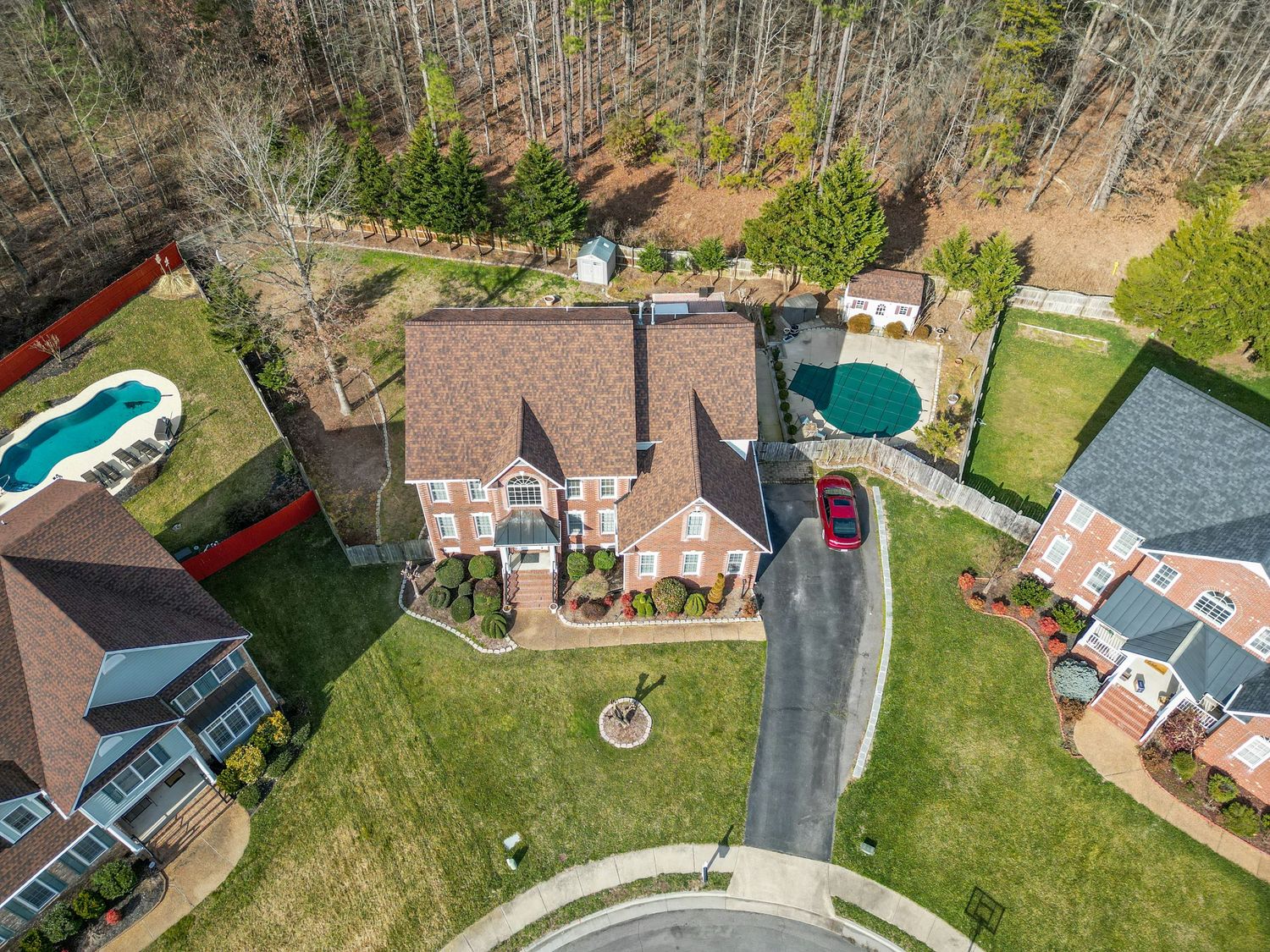 Aerial view of elegant brick colonial home with pool, circular driveway, manicured lawn, and wooded surroundings.