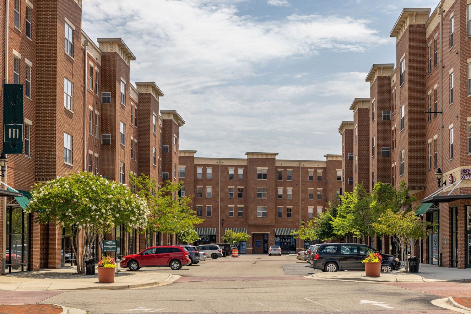 Symmetrical brick apartment complex with retail storefronts flanking a tree-lined urban courtyard on a cloudy day.
