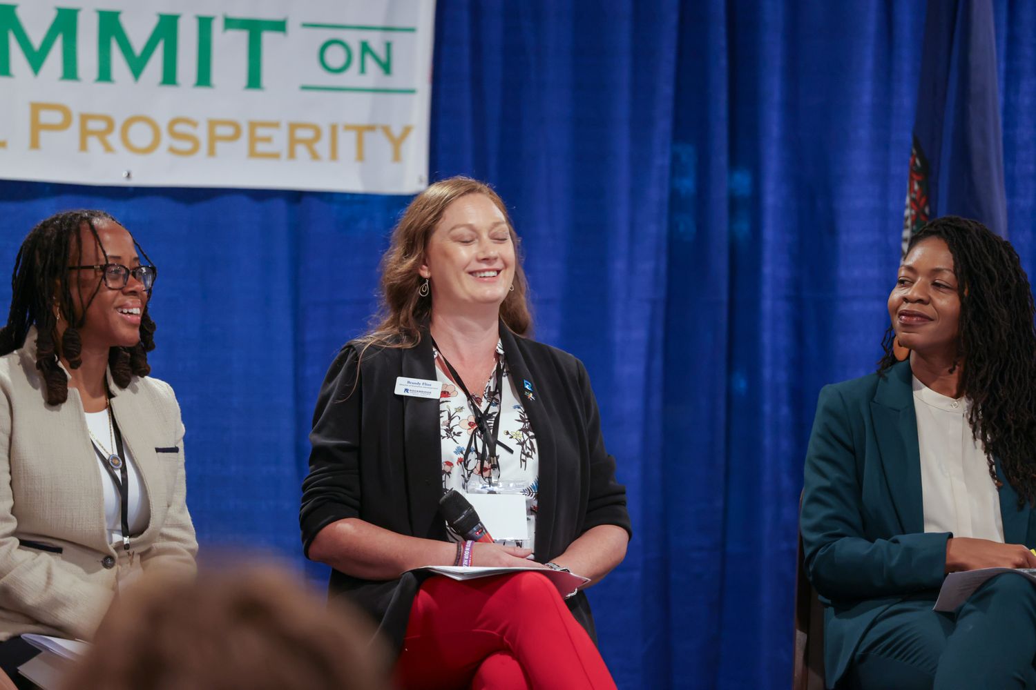 Three women smile and laugh together on stage at a Summit on Prosperity panel discussion event.