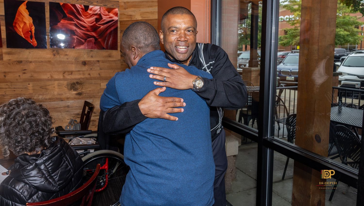 Two men share a warm hug inside a restaurant with wood-paneled walls and large windows, while a person in a wheelchair looks on.