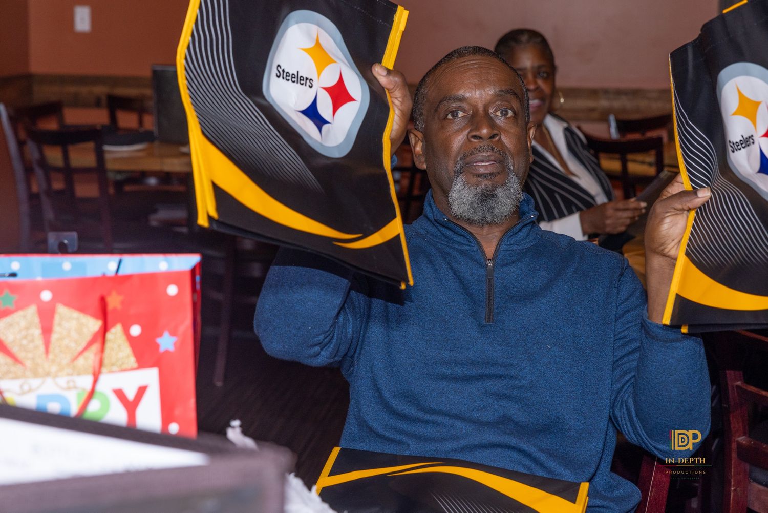 Smiling man in a blue sweater waves a black and gold Pittsburgh Steelers towel at a birthday party with a gift bag and guests nearby.
