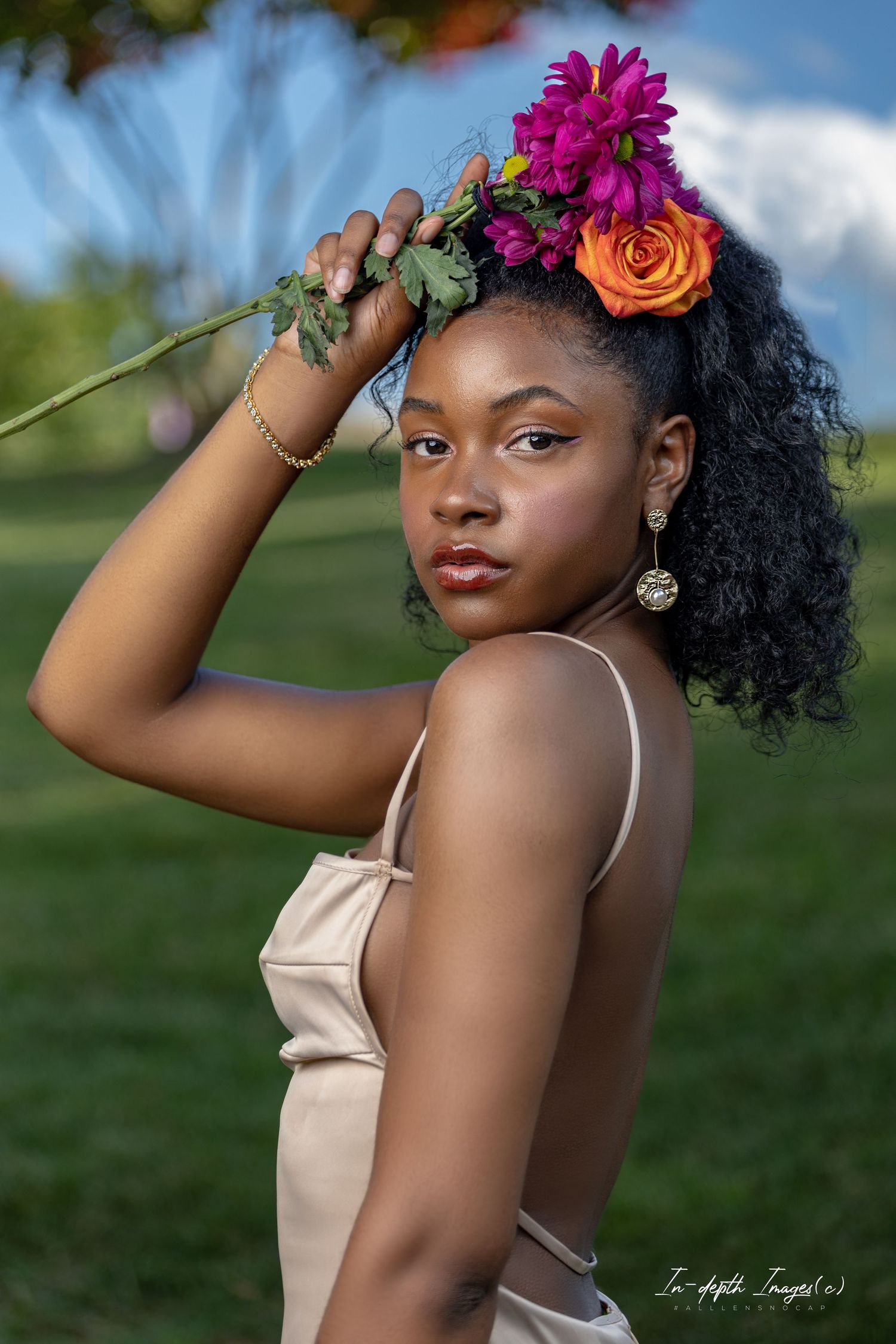 Young woman holding colorful flowers in hair poses outdoors in cream dress at golden hour.