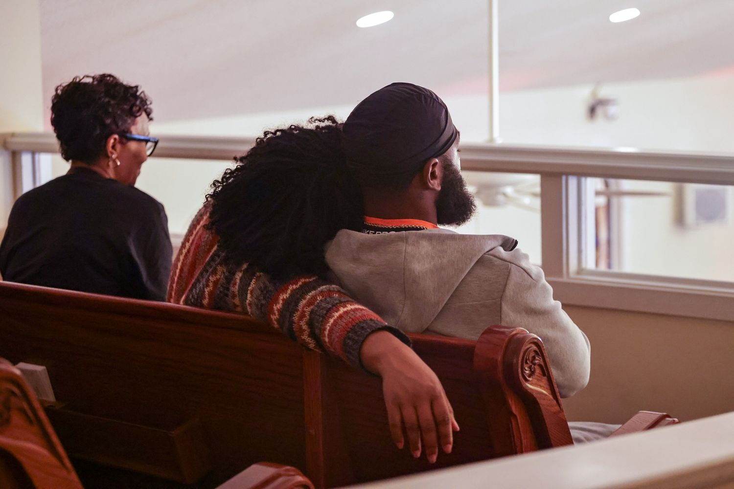 Two people sit close together on a wooden church pew, one resting an arm around the other, while a third person sits nearby.