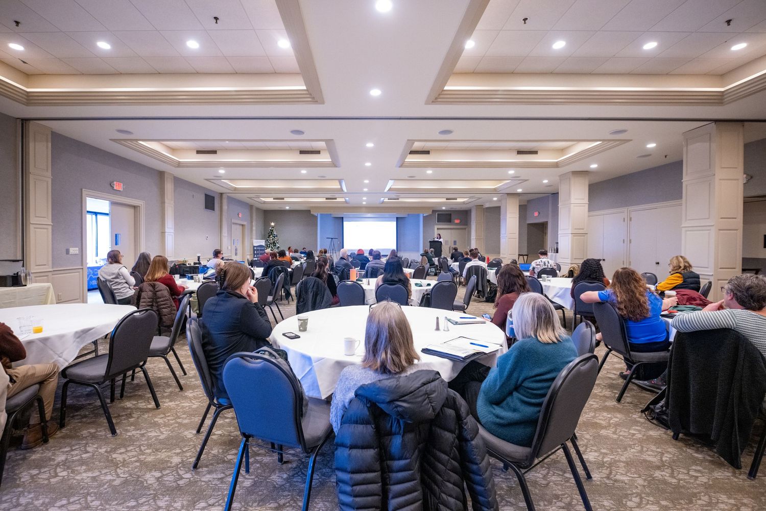 A large conference room filled with attendees seated at round tables during a professional seminar or business meeting.