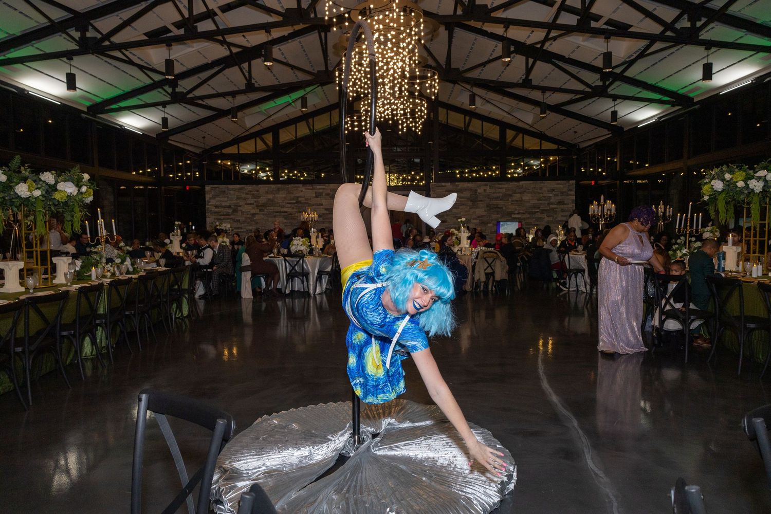 Acrobat in colorful costume performs aerial pole act at elegant wedding reception venue with chandeliers.