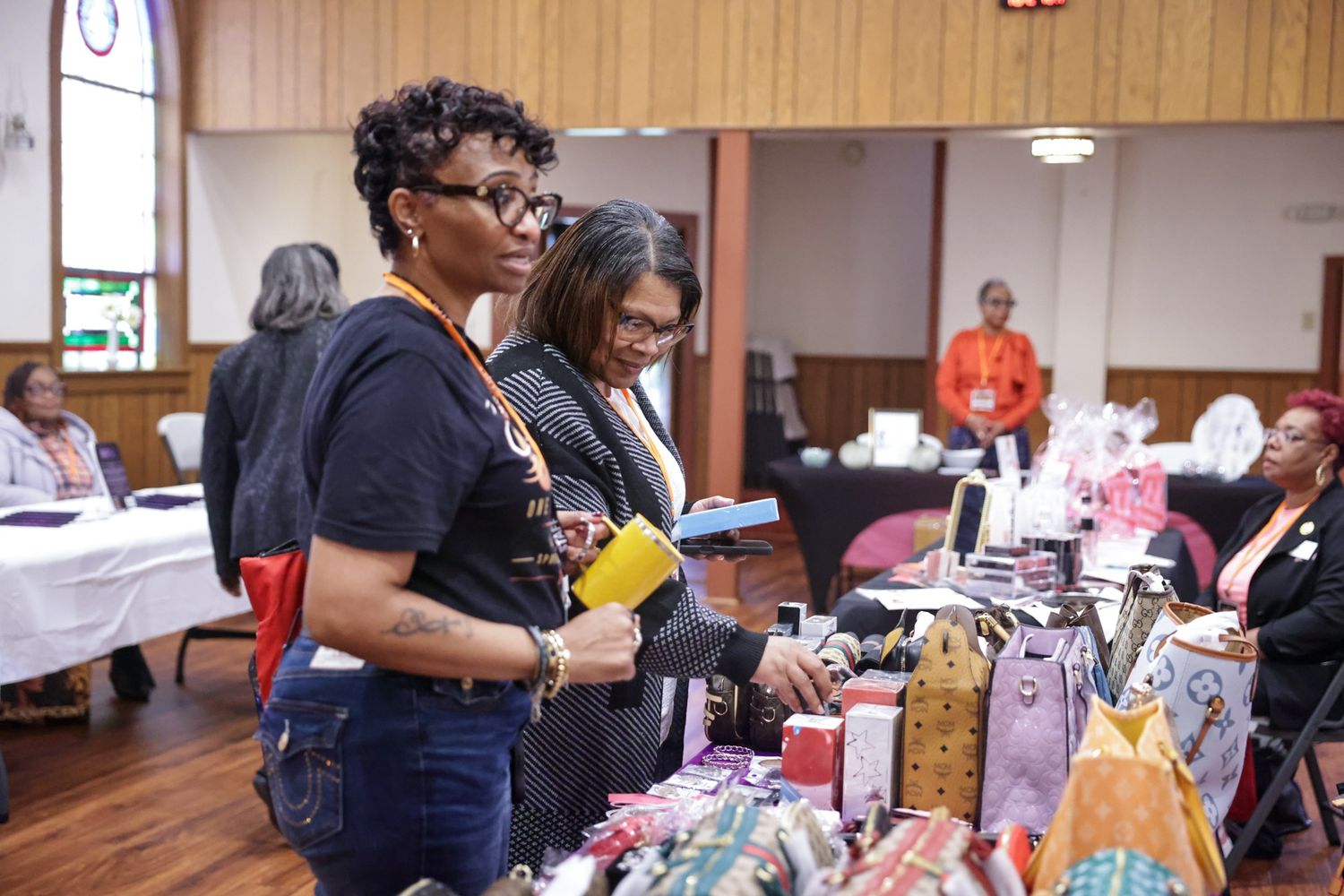 Two women browse handmade bags and crafts displayed on a vendor table at a community market inside a bright hall with wood-paneled walls.