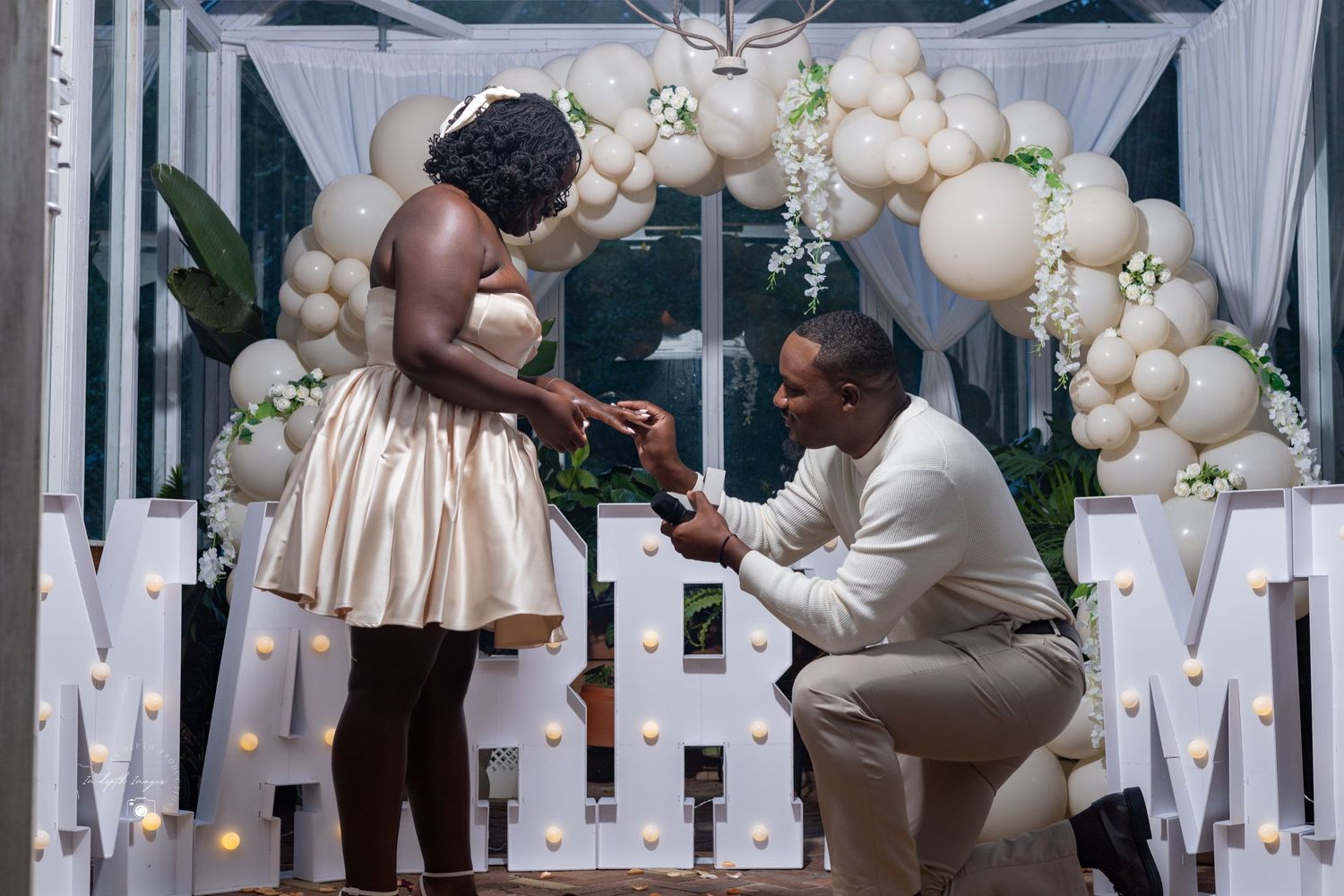Man proposes on one knee to woman in front of balloon arch and illuminated MARRY letters.