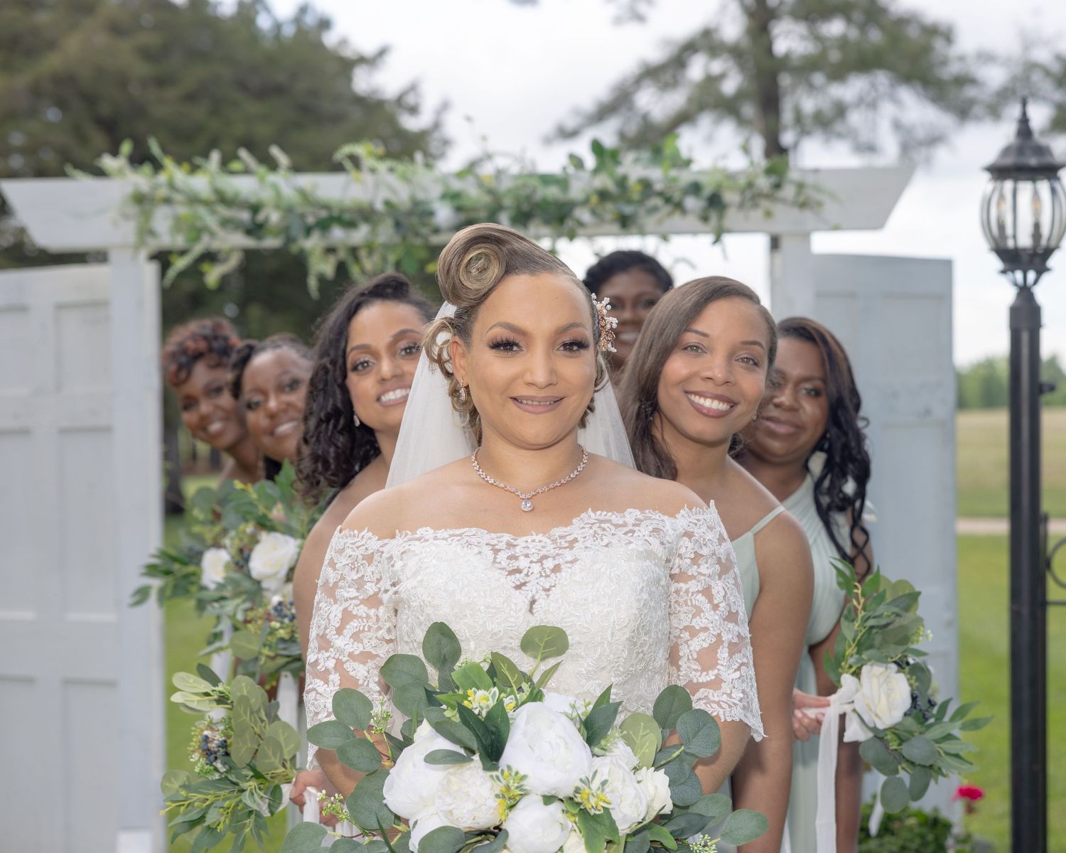 Bride leads bridesmaids through a white door arch adorned with greenery at outdoor ceremony.