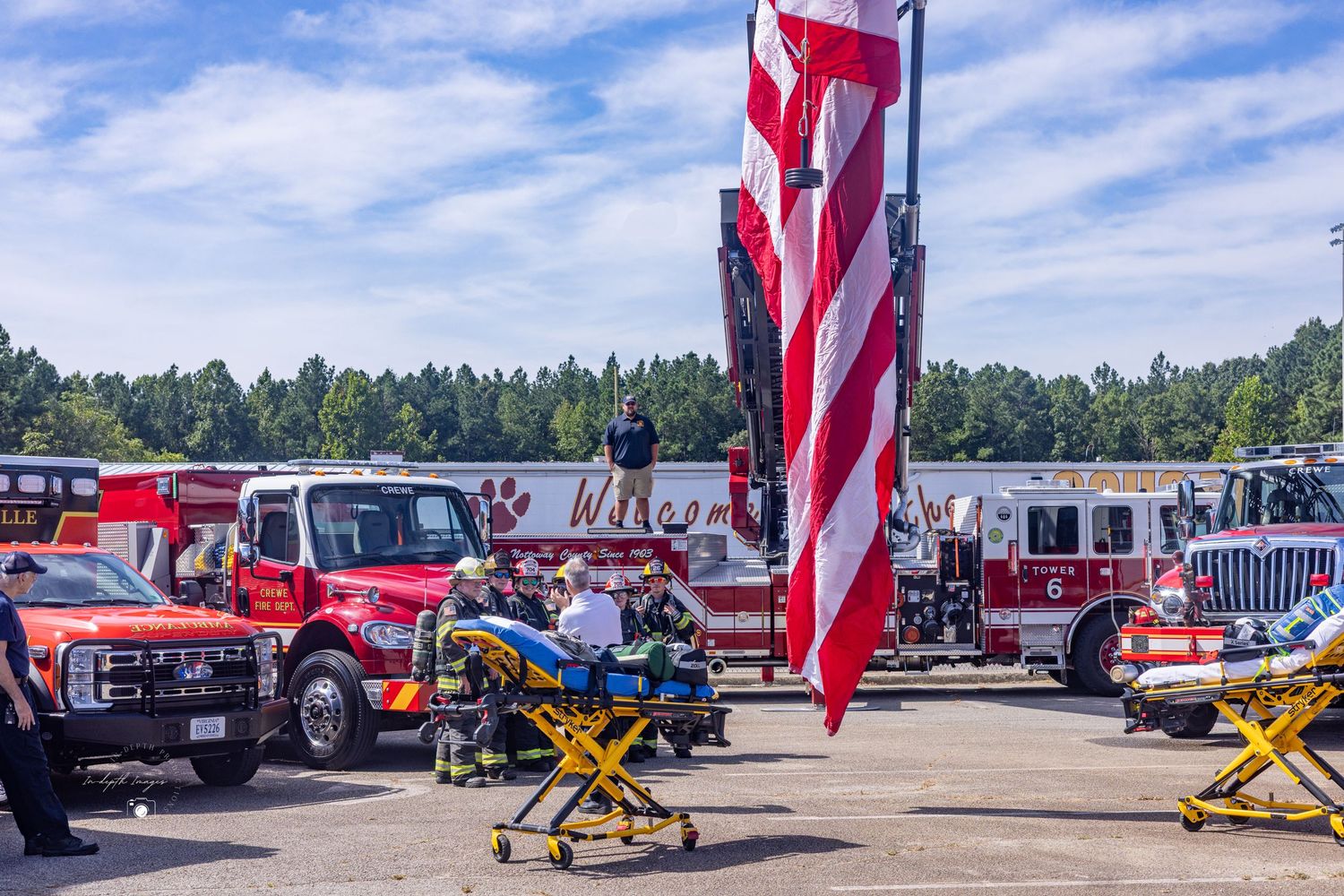 Firefighters and emergency responders gather near red fire trucks under a large American flag at an outdoor event.