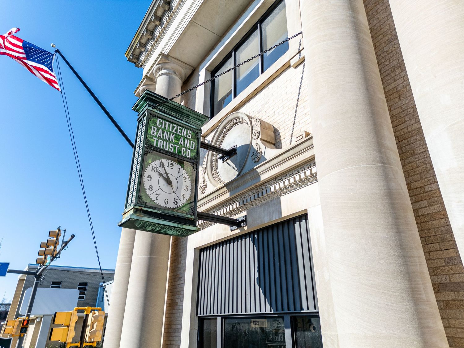 A green Citizens Bank and Trust street clock mounted outside a classic stone bank building with an American flag.