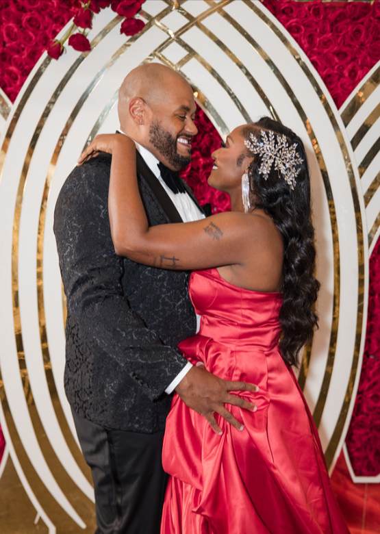 Couple in elegant formalwear embrace and laugh in front of a red rose and gold backdrop.