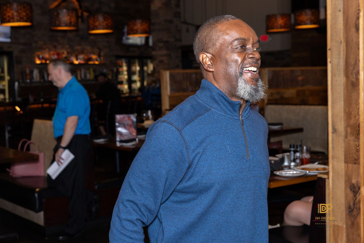Smiling bearded man in a blue quarter-zip pullover stands in a warmly lit restaurant with another man and dining tables behind him.