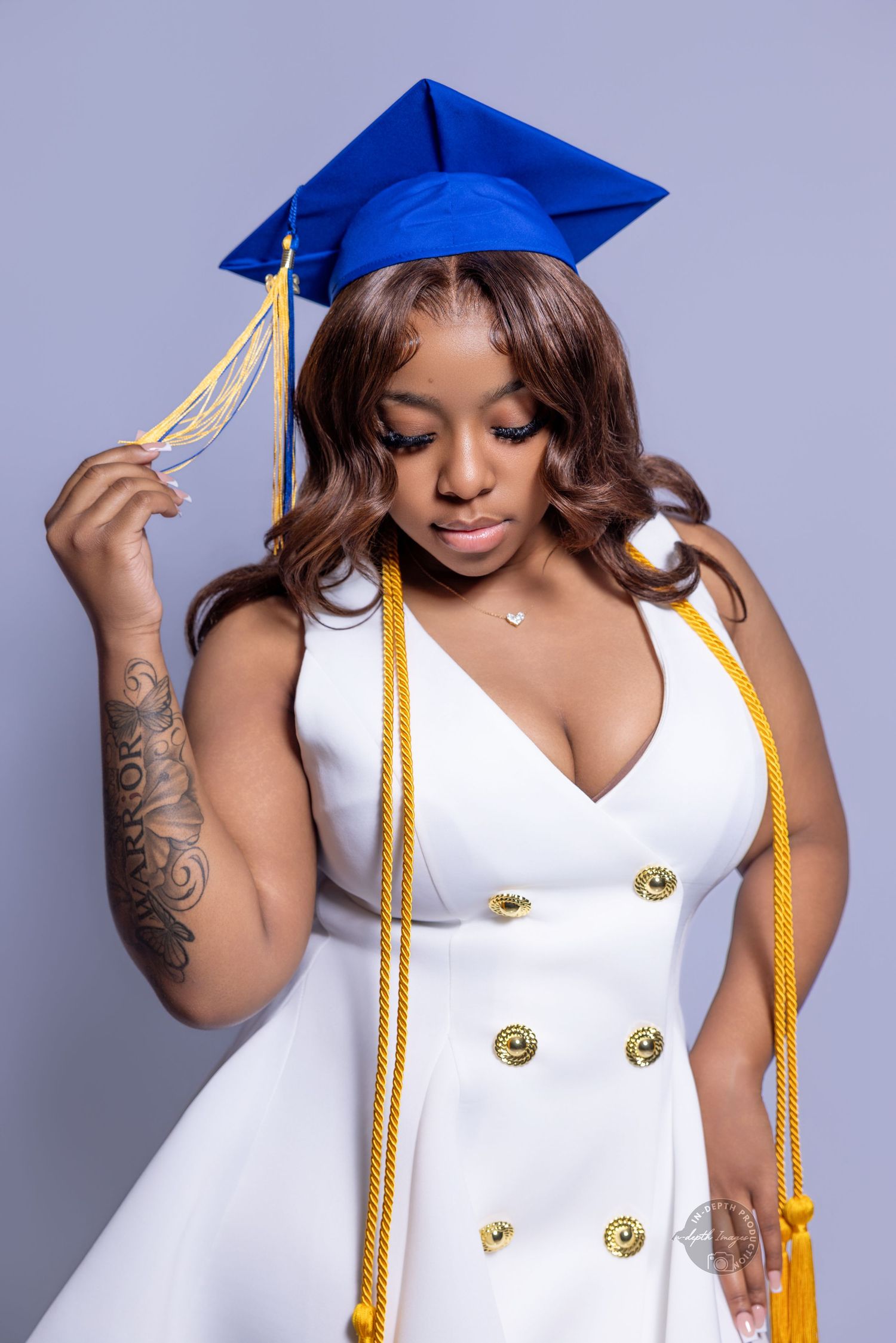 Young woman in white dress and blue graduation cap holding tassel, wearing gold honor cords for grad photo.