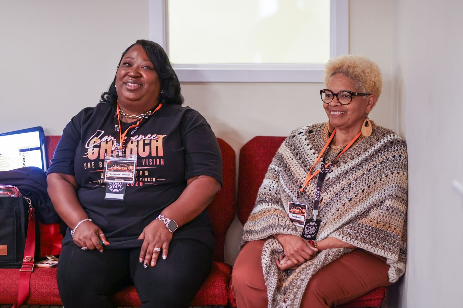 Two smiling women wearing conference lanyards sit on a red couch, one in a black graphic tee and the other in a patterned shawl.
