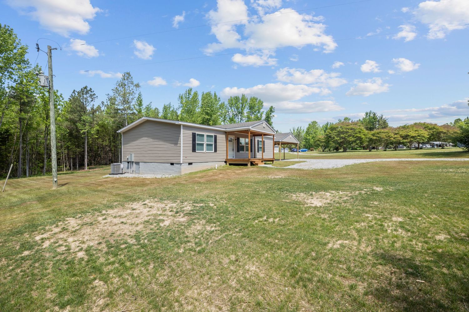 Small gray manufactured home with wooden deck surrounded by green lawn and tall trees on a clear day.