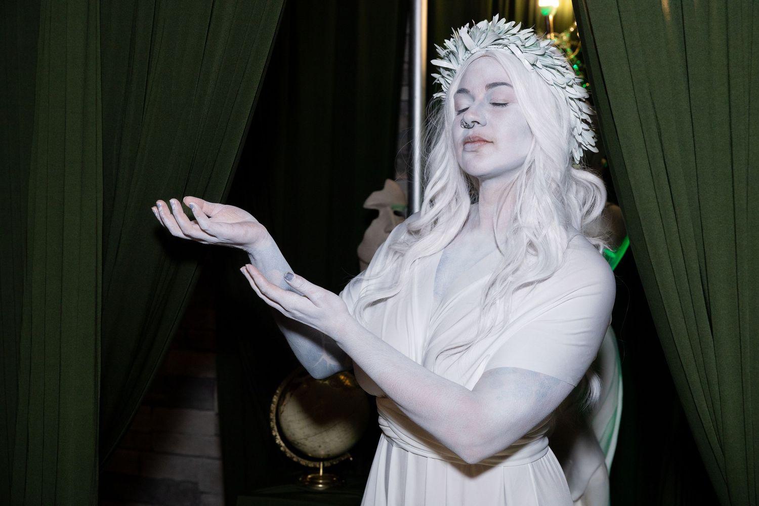 A woman in white costume and floral crown poses gracefully with outstretched hands against green curtains.