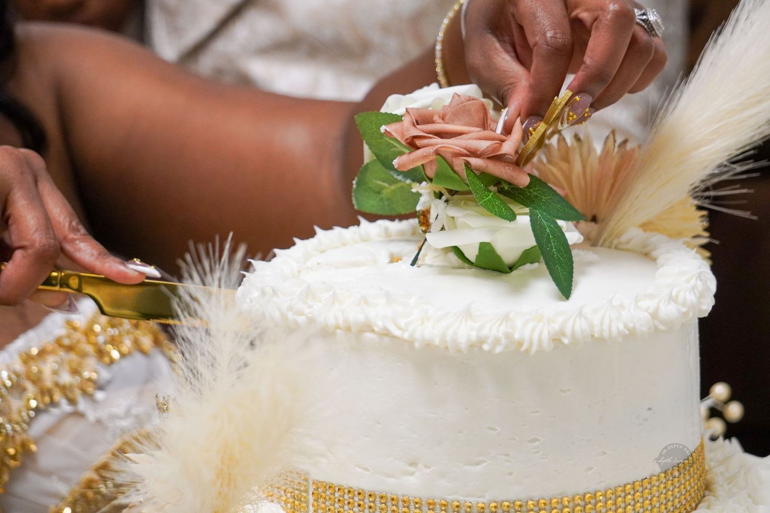 Two people cutting a white wedding cake decorated with pink flowers and greenery on a gold stand.