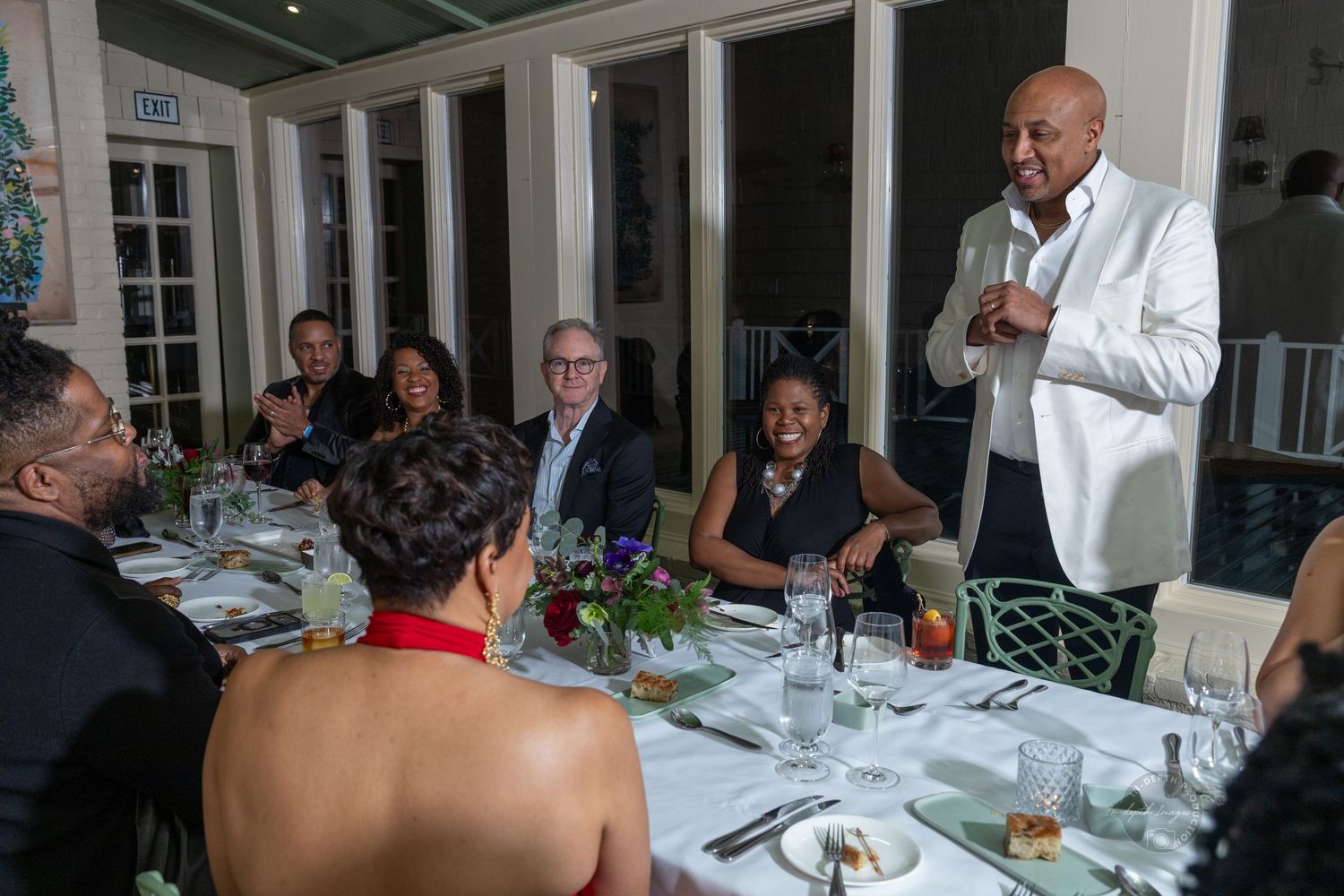 Elegant dinner party with guests seated at a decorated table while a man in a white jacket speaks.