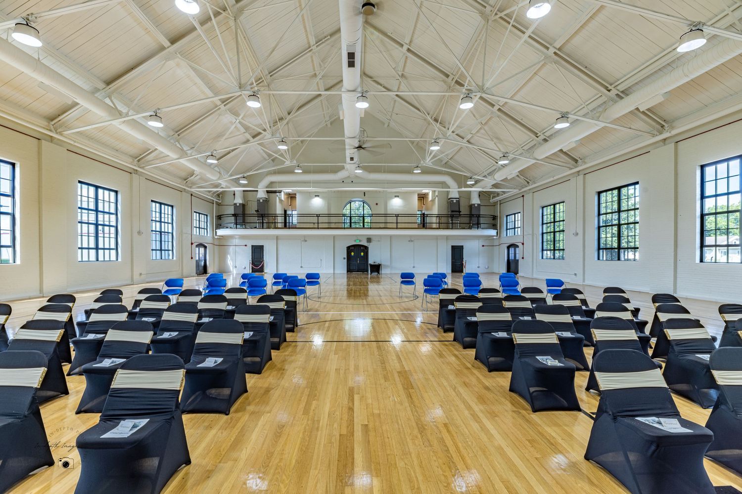 Spacious event hall with high ceilings, hardwood floors, rows of tables with black covers, and large windows.