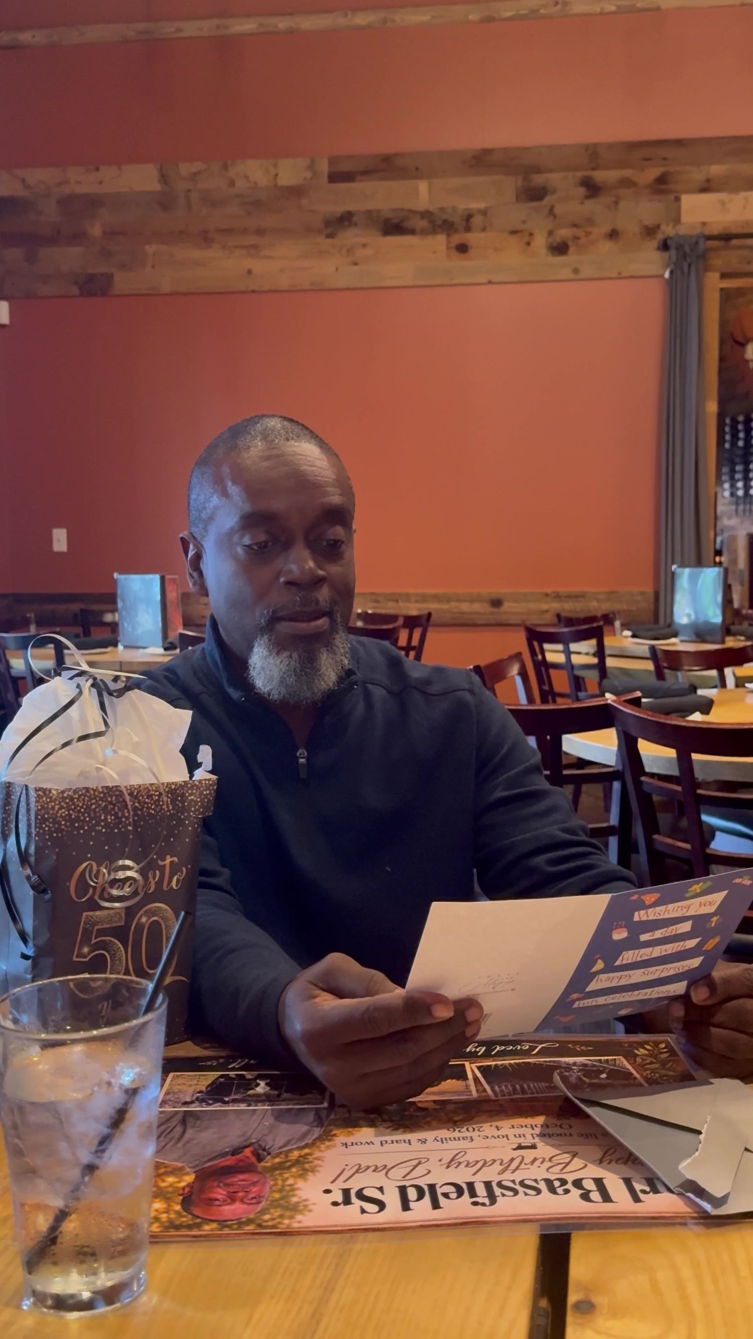 Man with a gray beard reads a greeting card at a wooden restaurant table beside a gold 50th birthday gift bag and a glass of ice water.