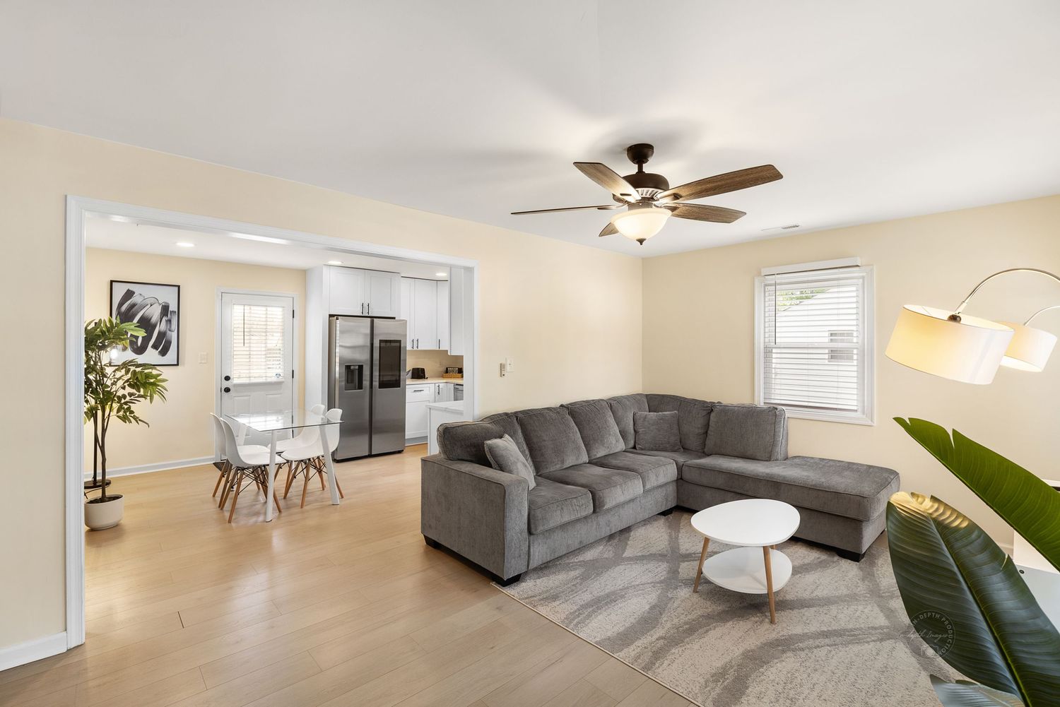 Bright living room with gray sectional sofa, ceiling fan, hardwood floors, and open kitchen with stainless steel appliances.