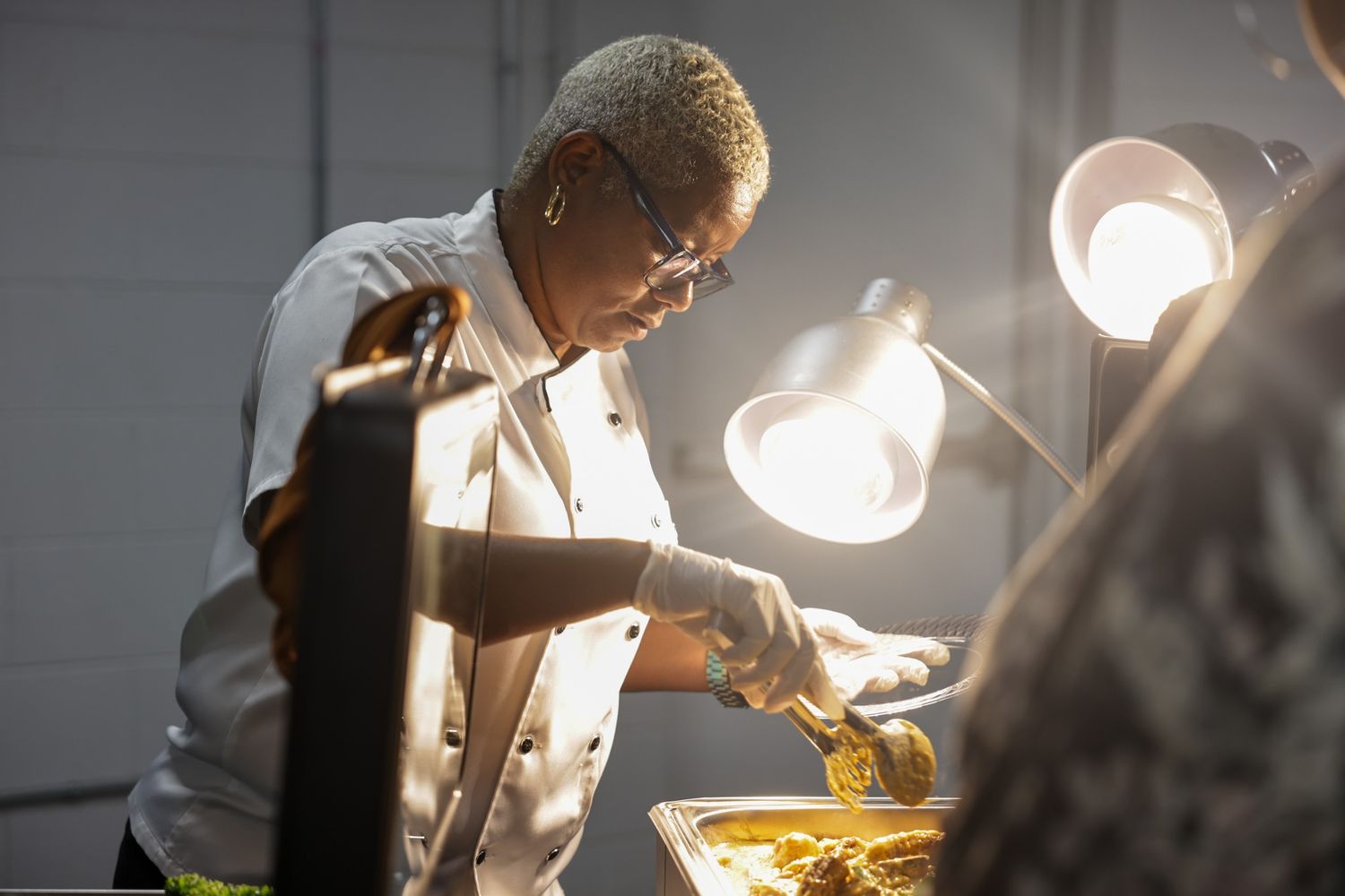 A chef in a white coat and gloves serves food from a warm buffet tray under glowing heat lamps.
