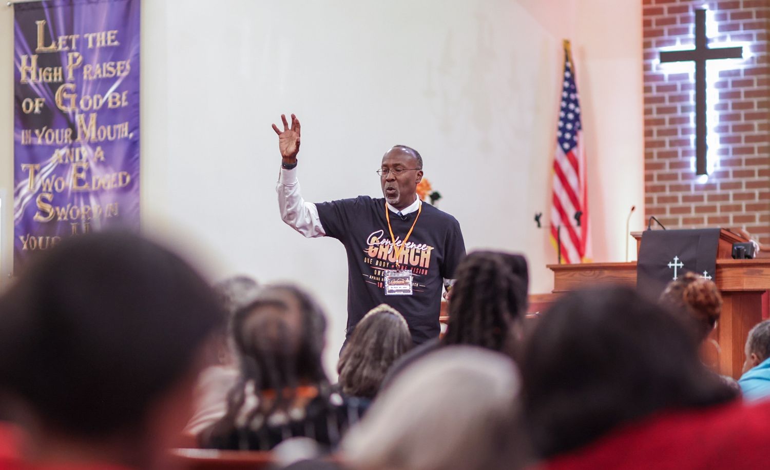 Speaker in a graphic sweatshirt raises one hand while addressing a seated congregation in a church with an American flag and lit cross.