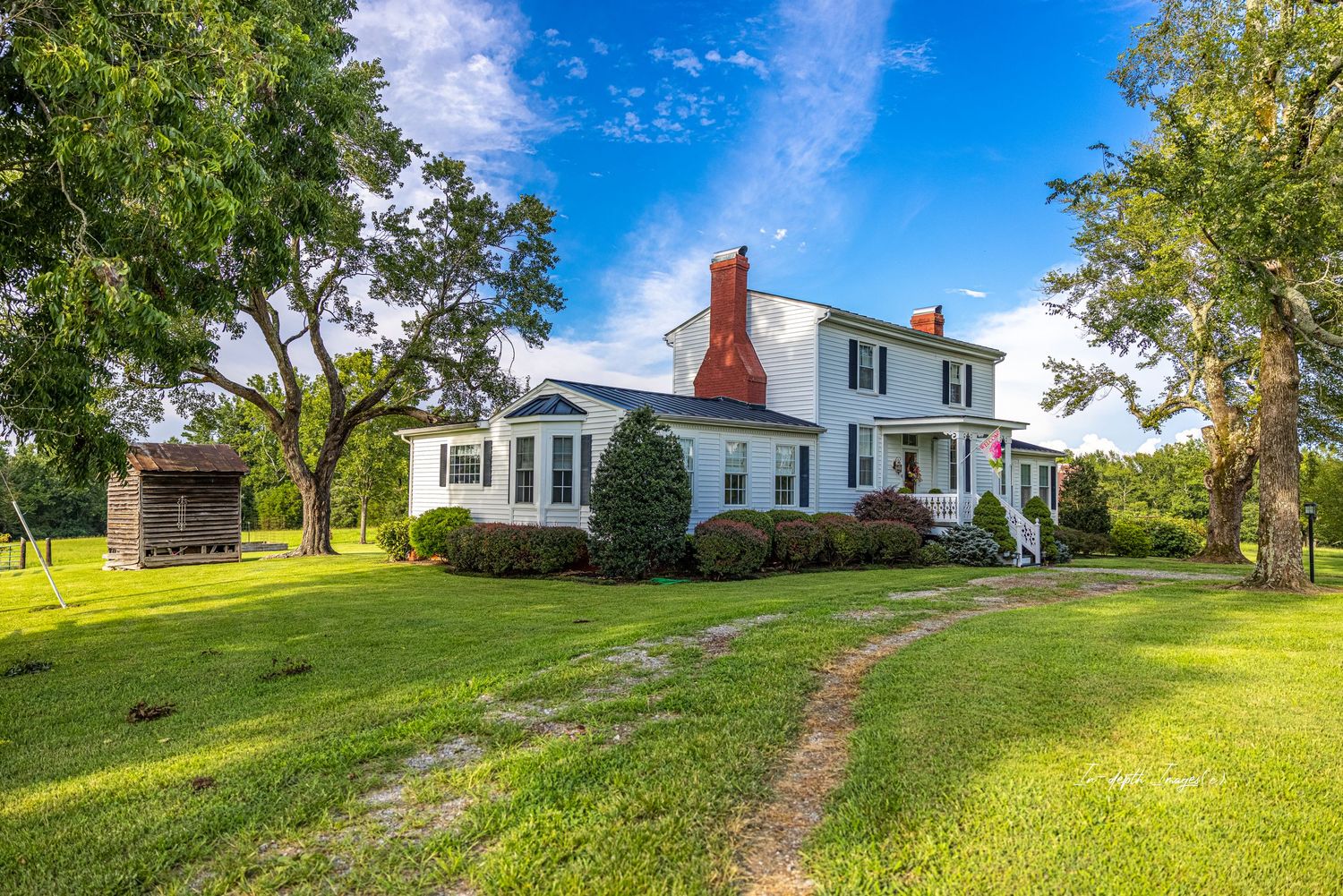 Classic white farmhouse with red chimney, mature trees, and lush green lawn on a bright summer day.