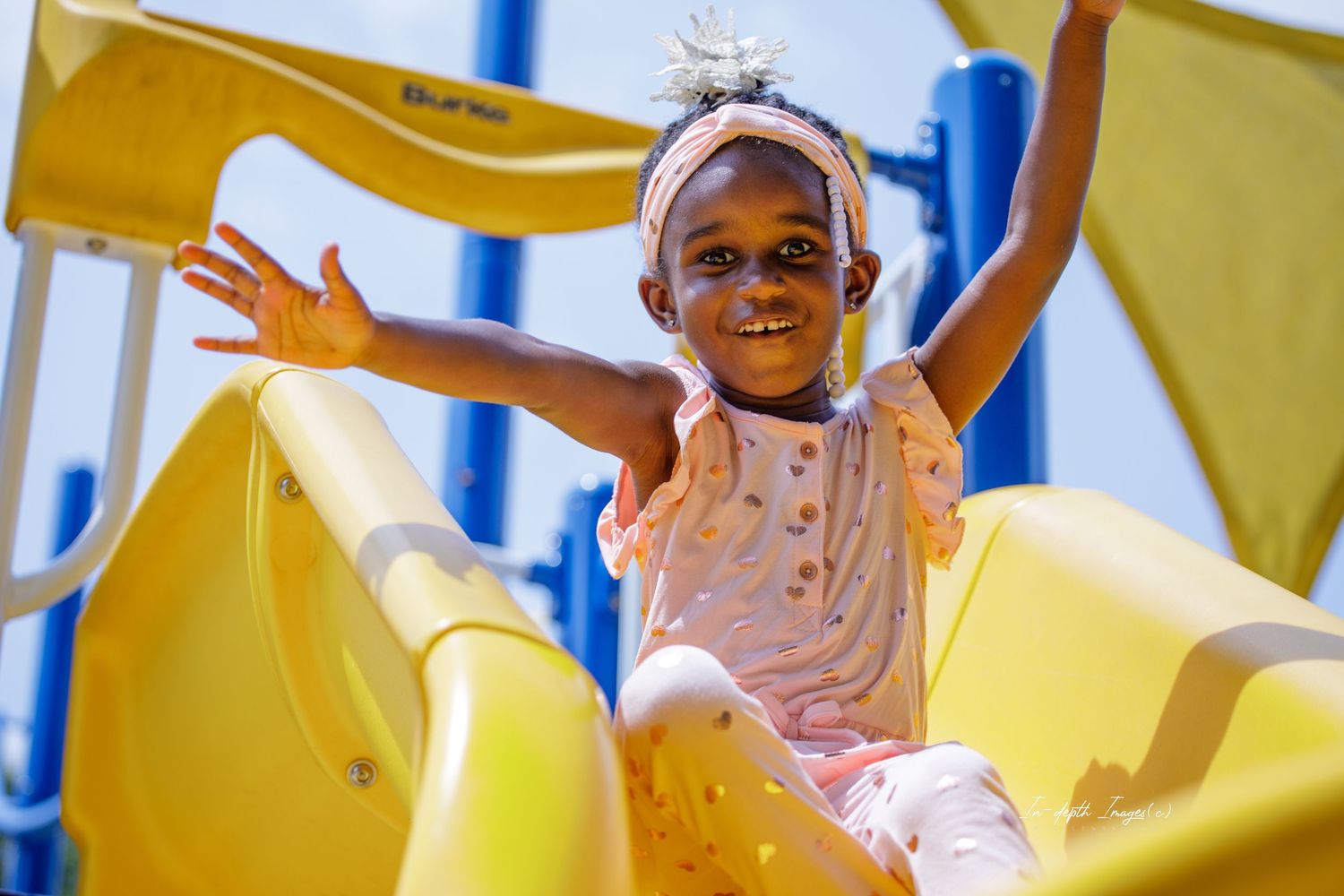 Happy young girl with arms raised sliding down yellow playground slide outdoors.
