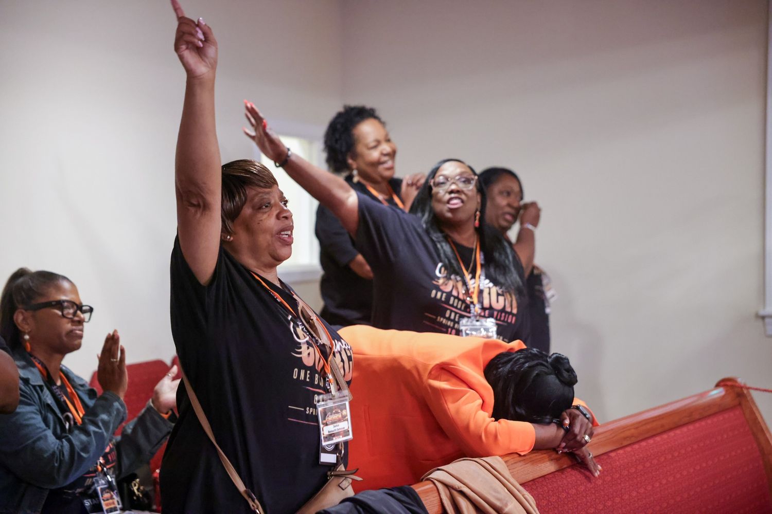 Group of women in black shirts and lanyards raise their arms in worship while a person in orange kneels in prayer at a church pew.