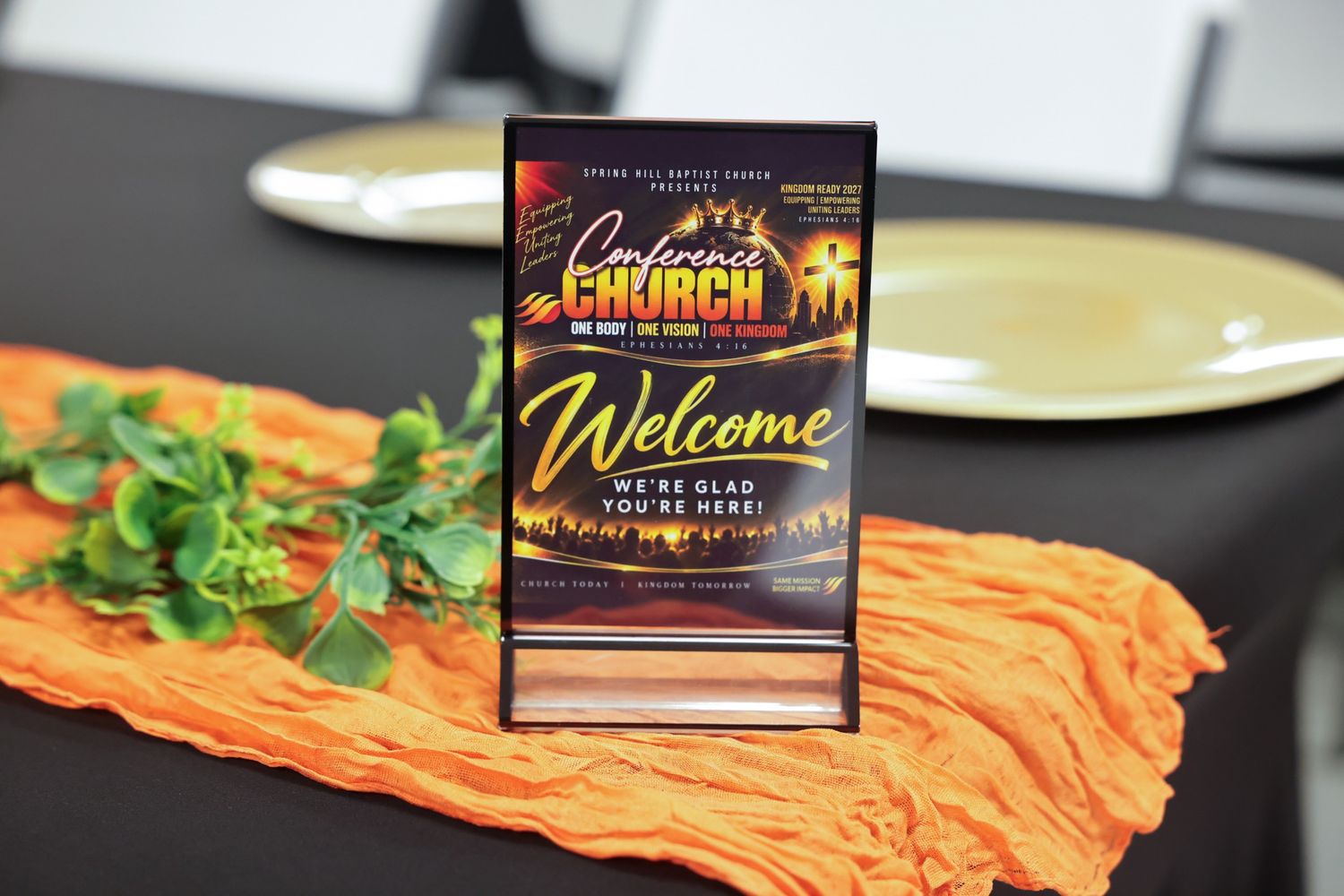 A Welcome church event flyer stands in a clear holder on a black table beside an orange cloth runner, green leaves, and gold plates.