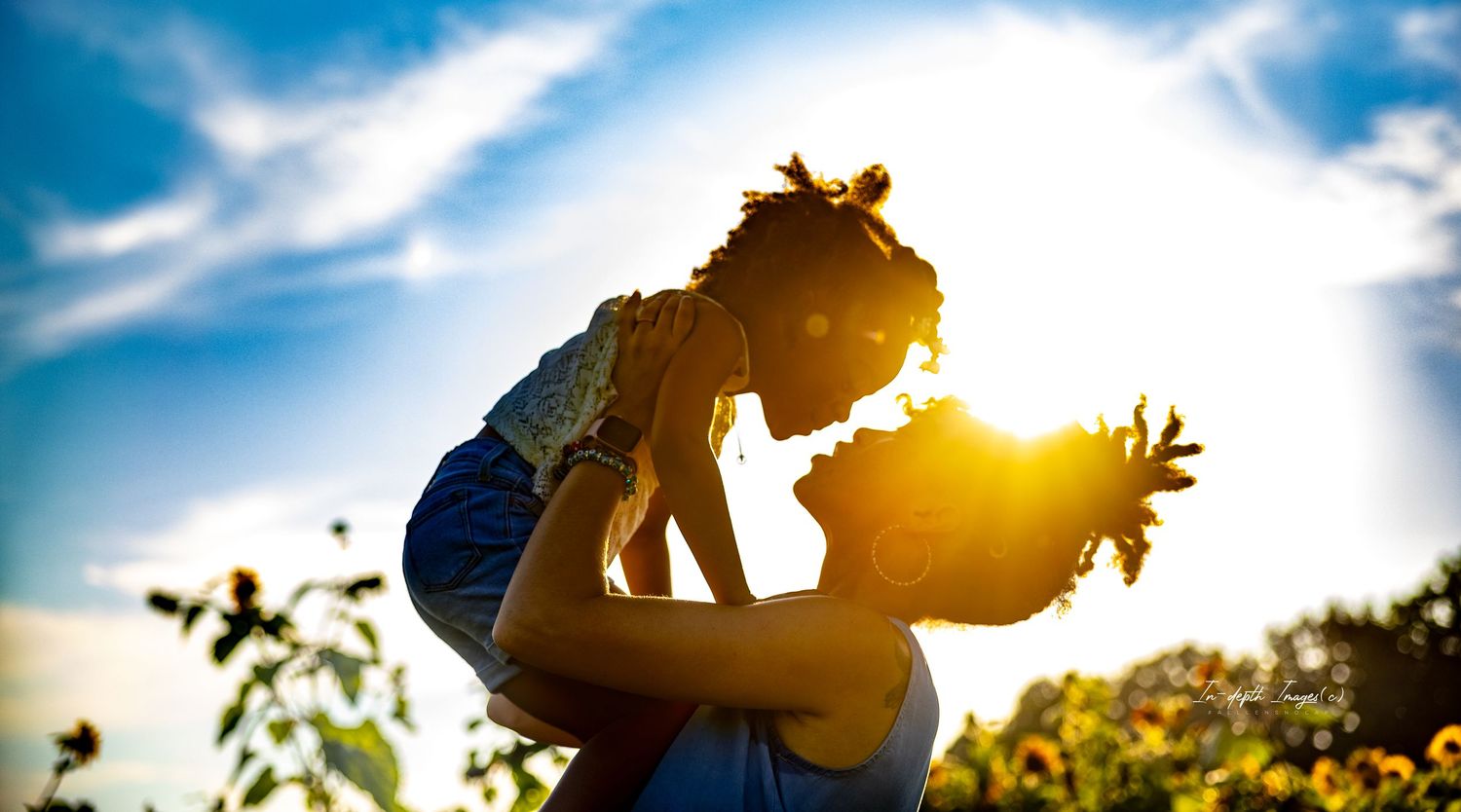 A mother joyfully lifts her child against a golden sunset sky in a sunflower field.