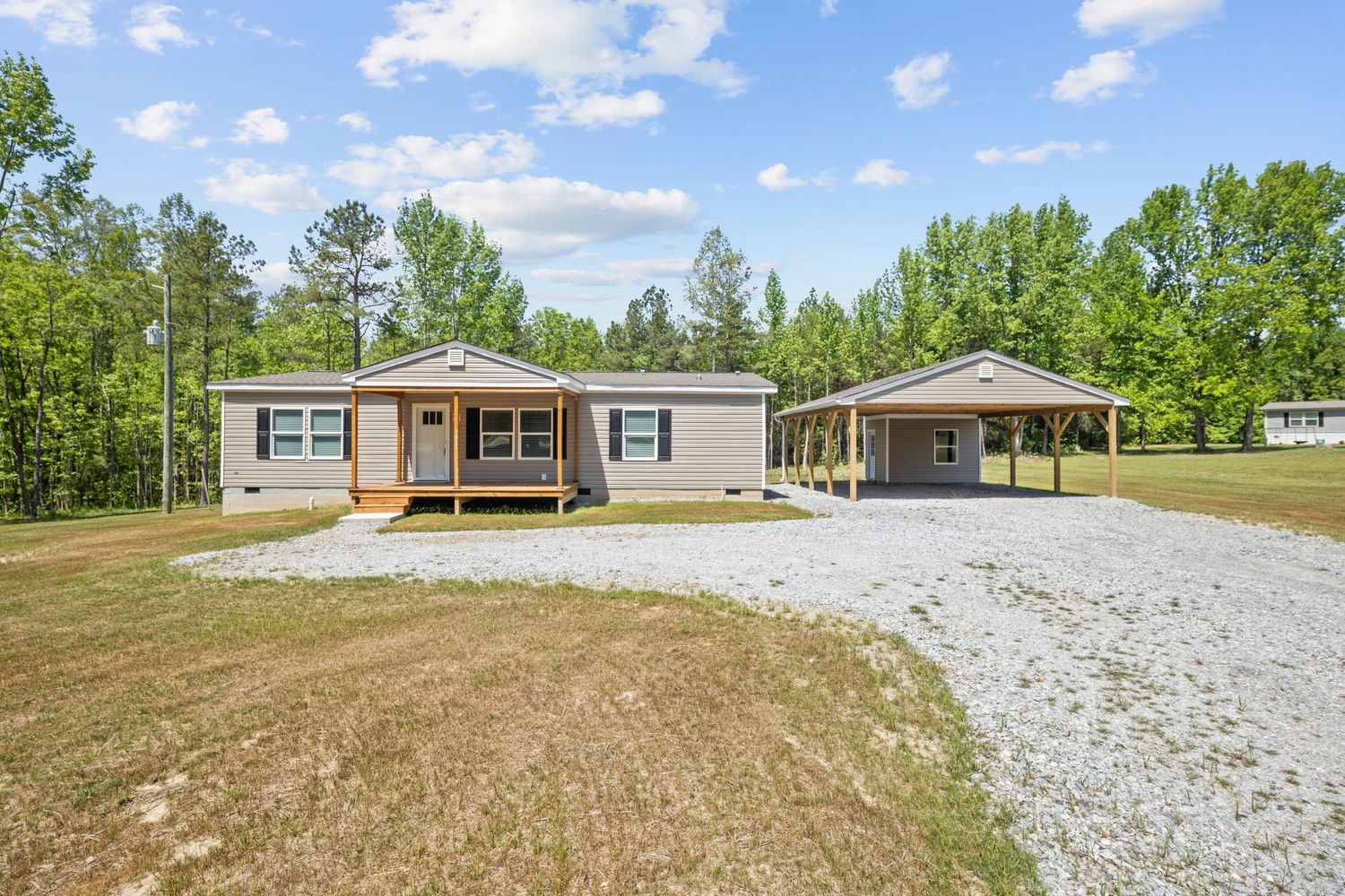 Single-story manufactured home with covered porch, attached carport, gravel driveway, and lush green lawn surrounded by tall trees.