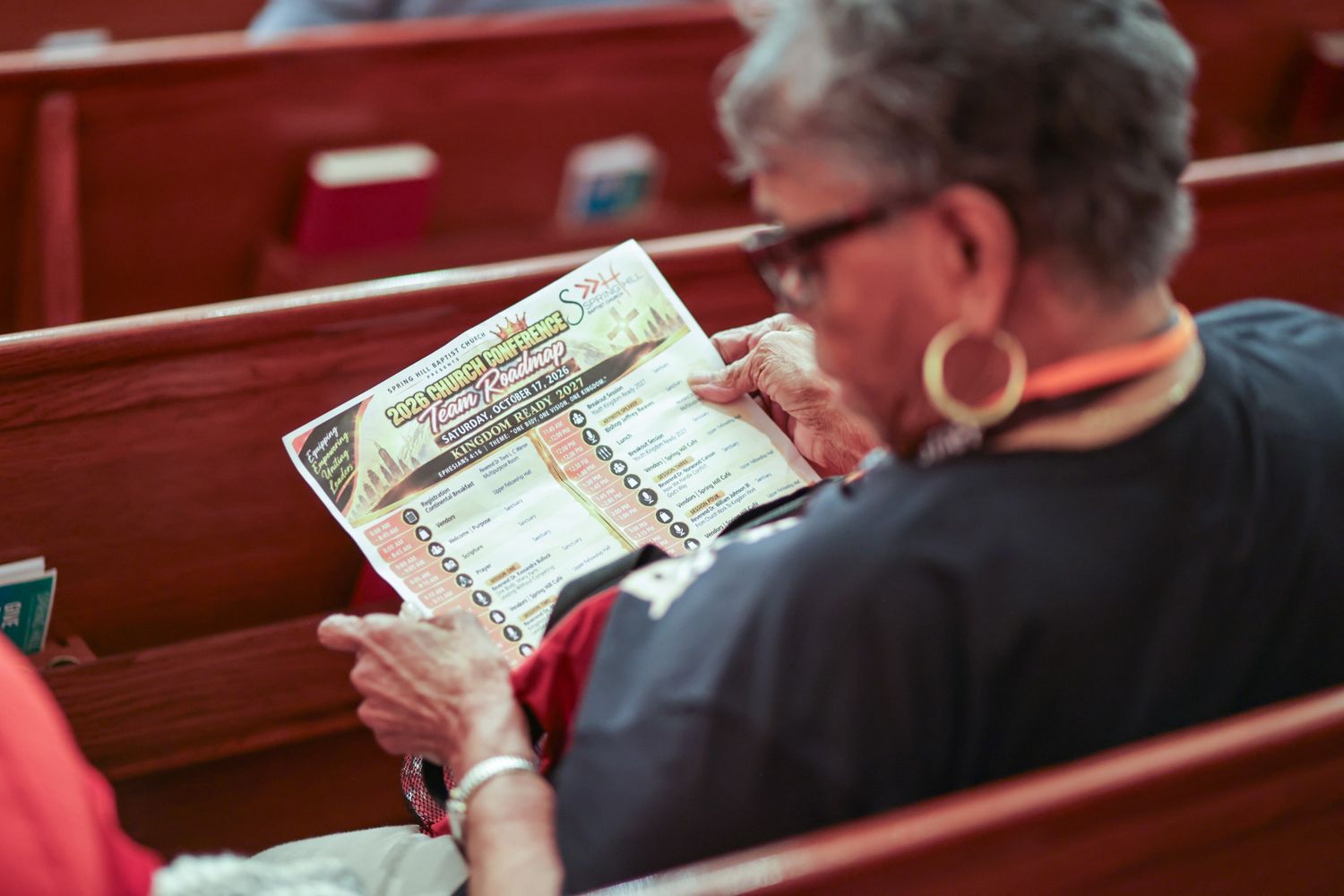 Congregation member in glasses and large hoop earrings studies a printed event program while seated in a wooden church pew.