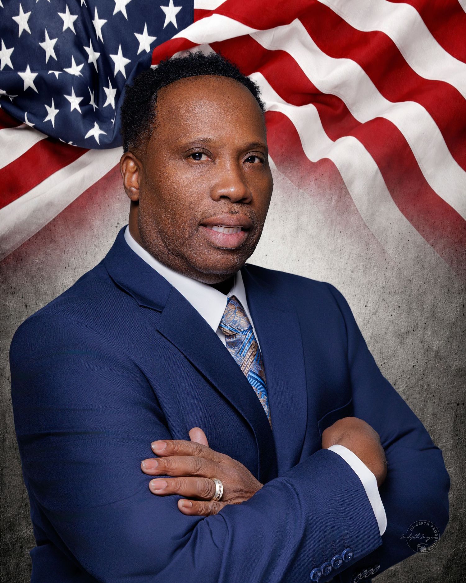 Professional man in blue suit with arms crossed poses before an American flag backdrop in a formal portrait.