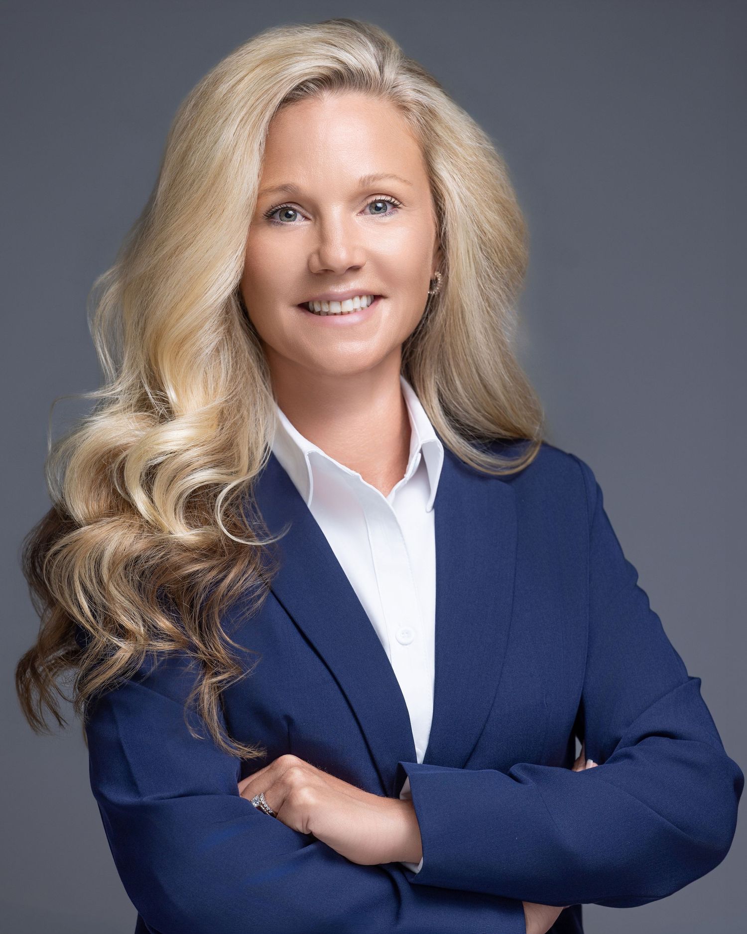 Professional woman in navy blazer with blonde hair, arms crossed, smiling confidently against gray background.