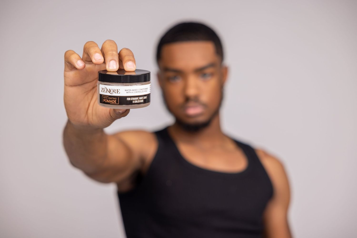 Young Black man in black tank top holding a small jar of Zenore grooming product toward the camera.