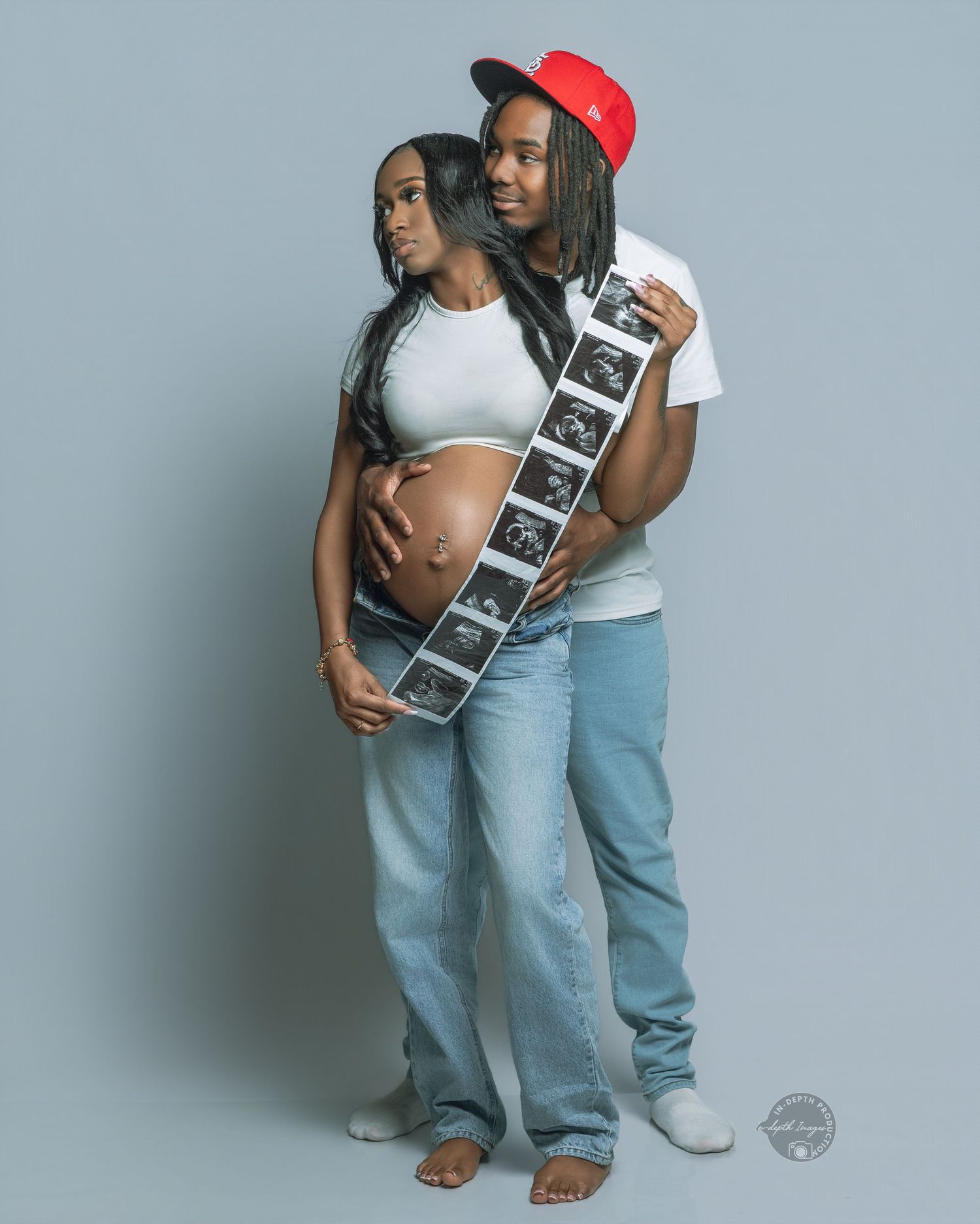 Pregnant couple posing with ultrasound strip, woman in crop top showing baby bump, man in red cap embracing her from behind.