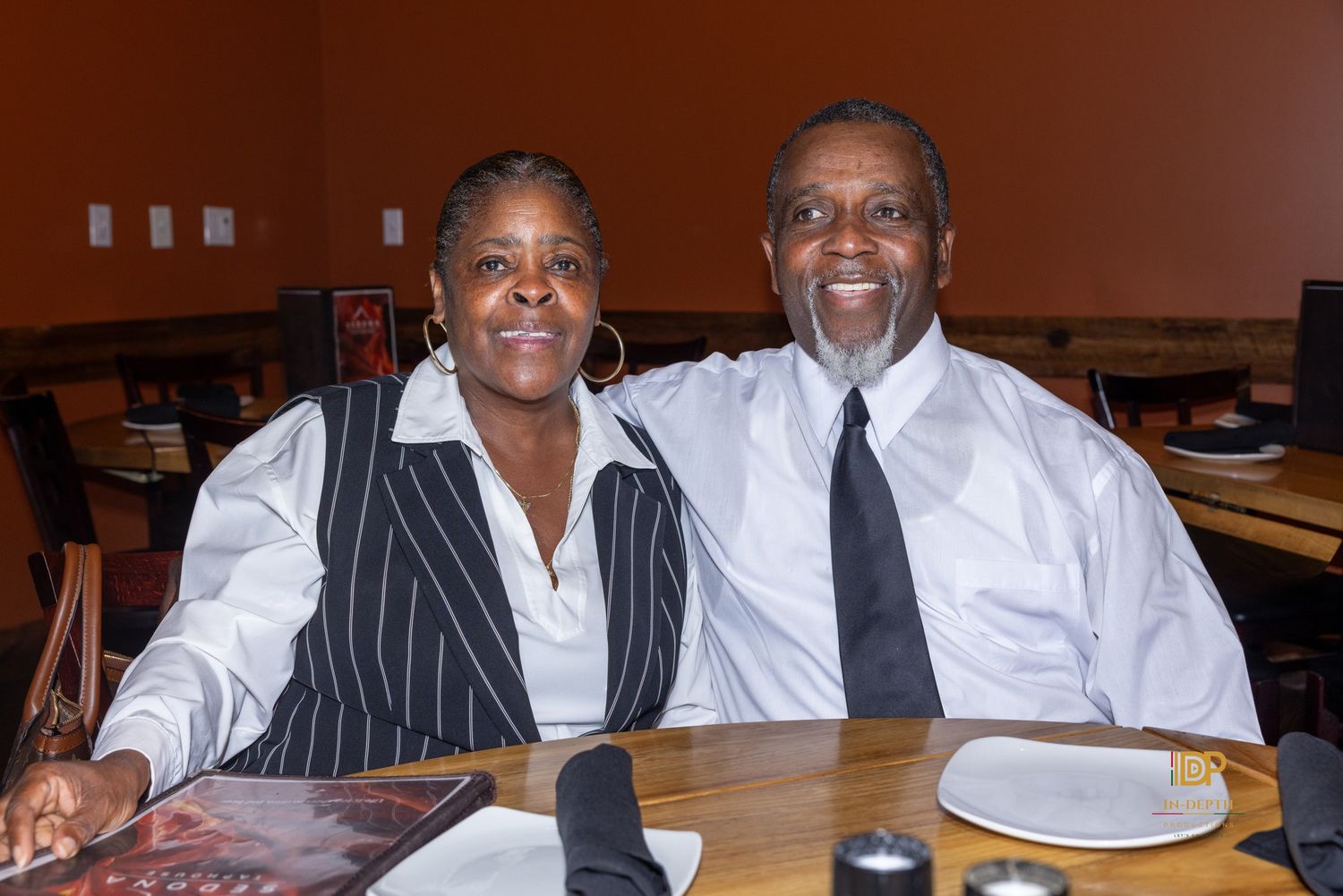 Smiling man and woman in dressy attire sit side by side at a wooden restaurant table with napkins, plates, and a menu in front of them.