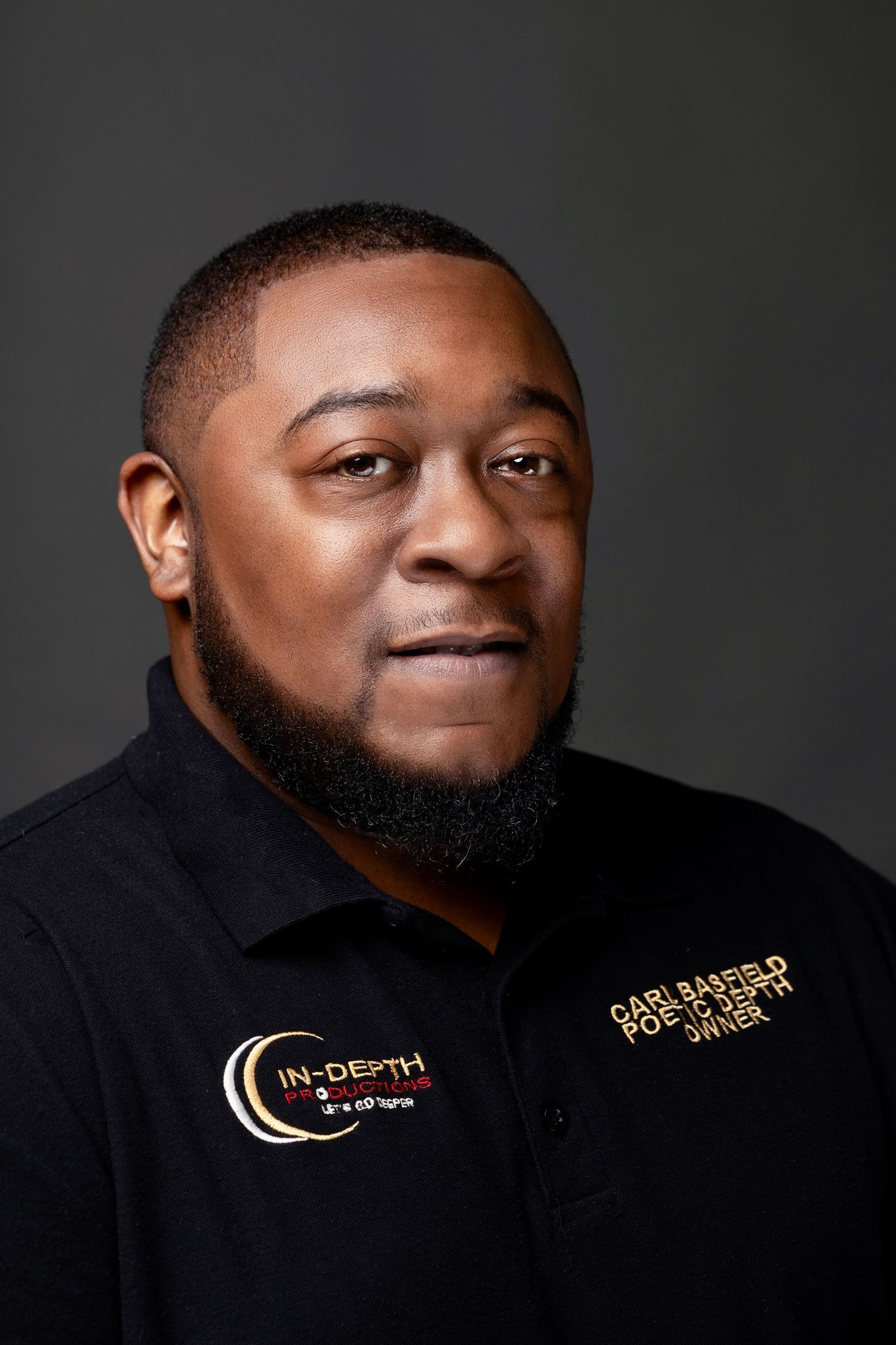 Professional headshot of a bearded man in a black polo shirt with company logo on gray background.