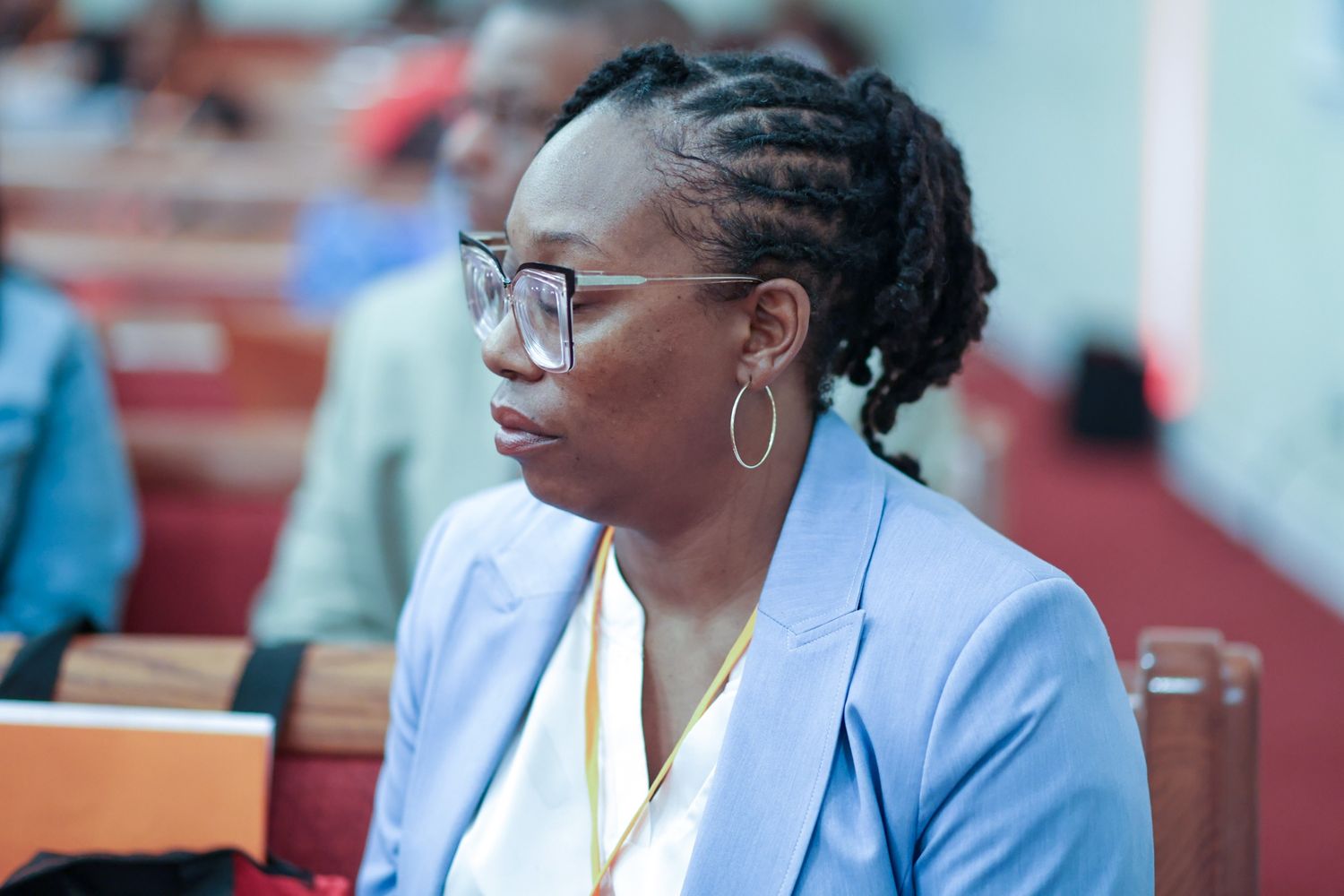 Woman with braided hair and glasses, wearing a light blue blazer, listens attentively while seated among attendees in a church pew.