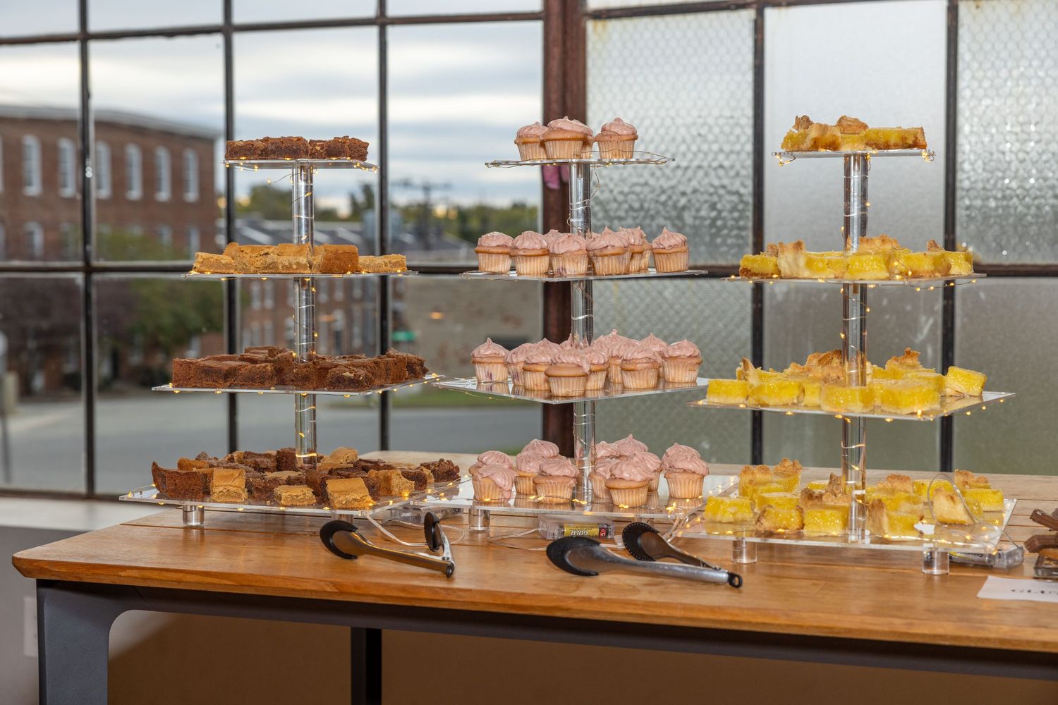 Elegant multi-tiered dessert display with cupcakes, pastries, and cakes on a wooden table by large windows.
