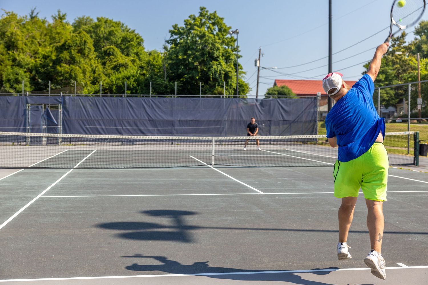 Two tennis players competing on an outdoor hard court on a sunny day, one mid-serve in blue shirt and yellow shorts.